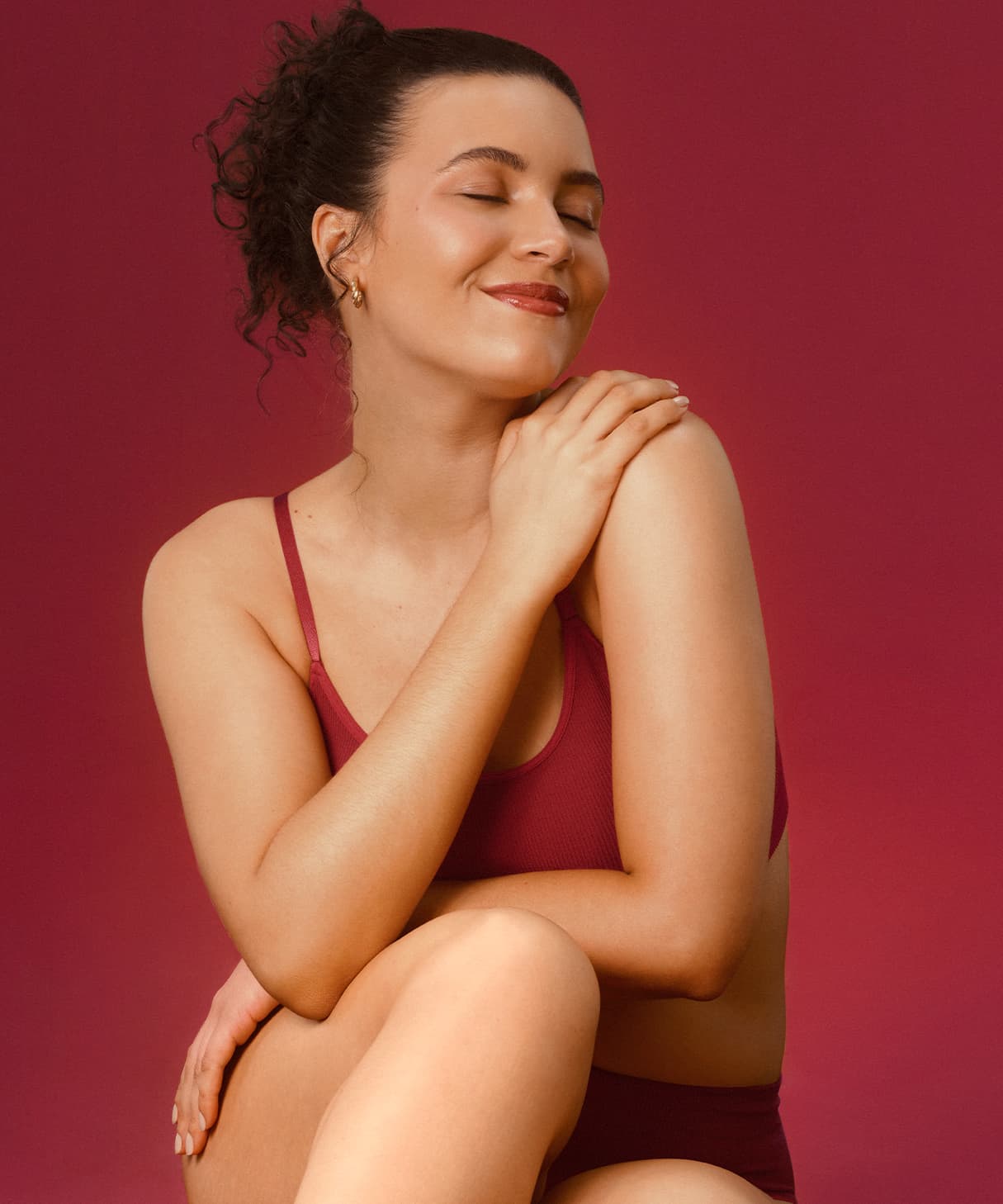 Imagem de mulher de cabelos cacheados usando top vermelho, sentada com os braços cruzados em um fundo vermelho. Ela espalha um pouco de loção hidratante corporal no ombro. Está sorrindo e de olhos fechados.