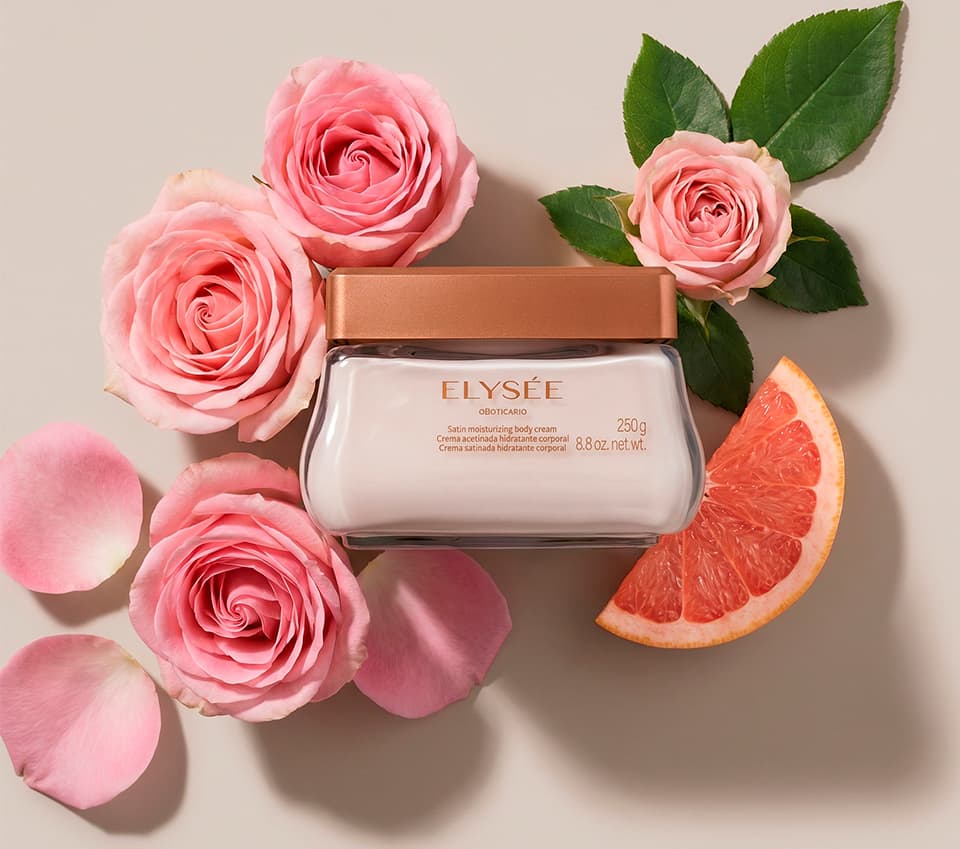 Imagem do pote de creme corporal da marca Elysée em primeiro plano, rodeado por rosas cor de rosa e fatia de toranja sobre fundo bege claro. O ambiente sugere um contexto de feminilidade, delicadeza e frescor.