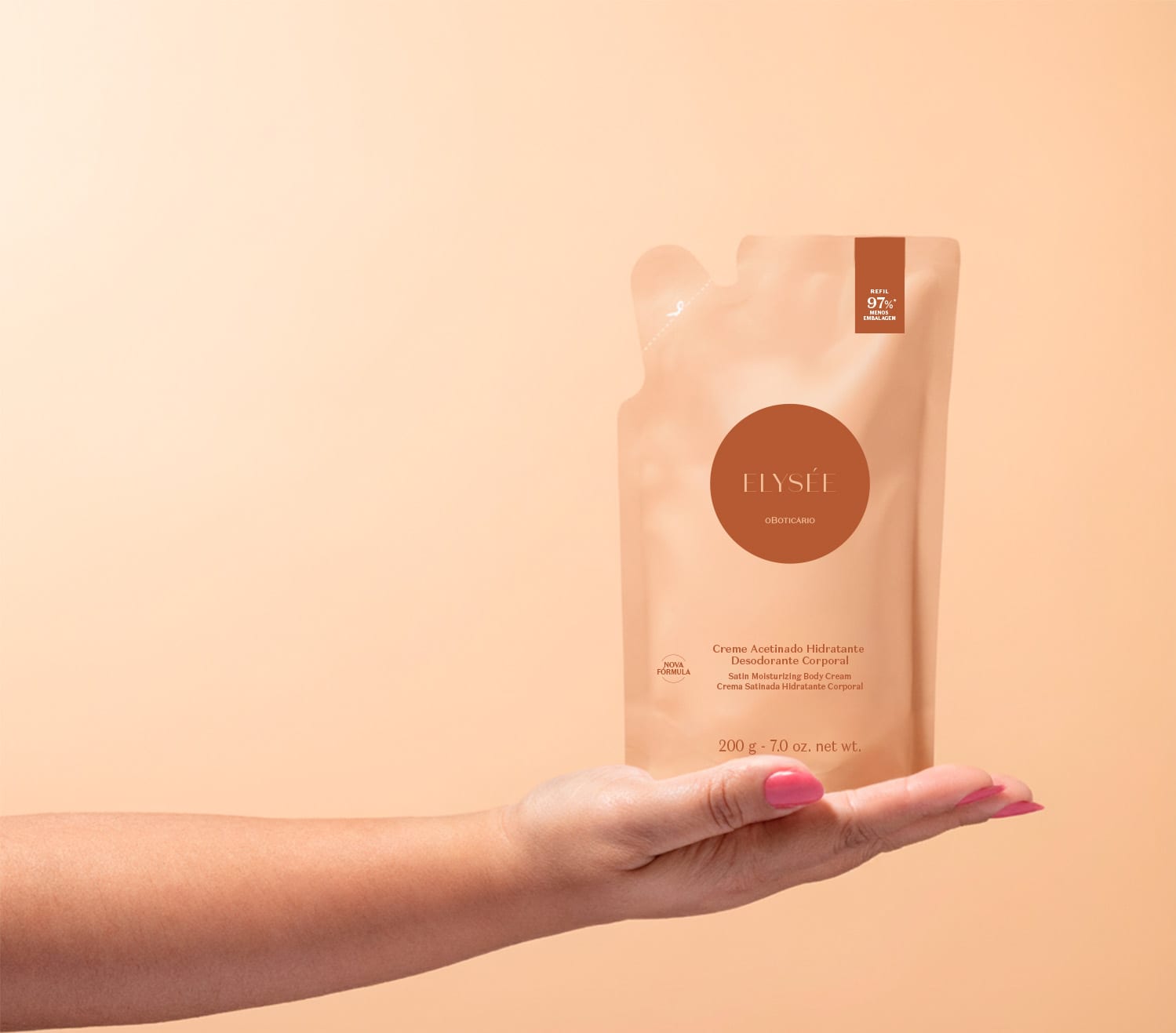 Imagem de uma mão segurando um refil de creme hidratante desodorante corporal da marca Elysée, em embalagem de sachê com predomínio de tons de laranja e marrom, contendo informações sobre a fórmula e o volume do produto. O fundo é em tons de rosa.