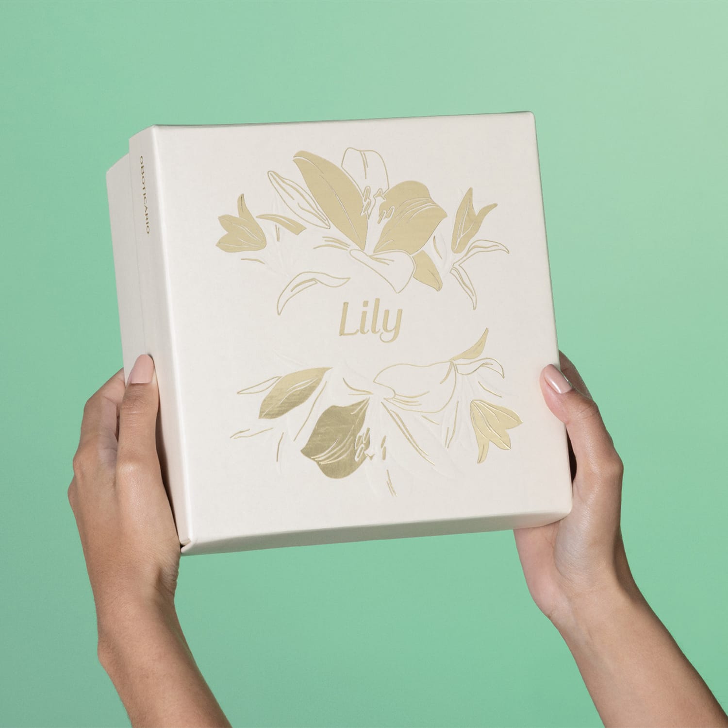 Imagem de uma caixa branca com ilustração dourada de flores, sobre fundo verde claro, segura por mãos femininas.