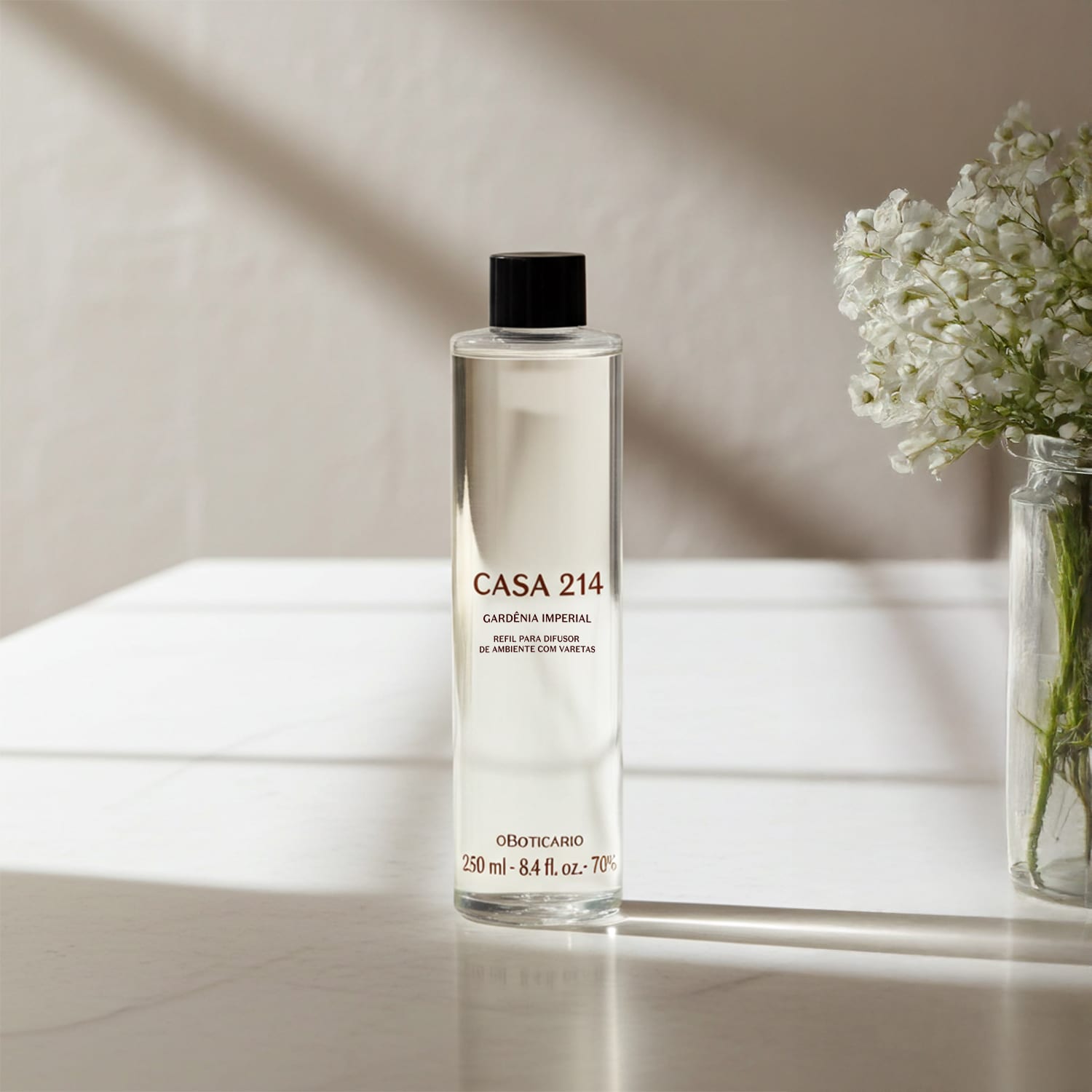 Imagem de um frasco refil com a fragrância Gardênia Imperial para difusor de ambiente da marca Casa 214 em embalagem transparente e tampa preta, posicionado sobre superfície clara com vaso de flores brancas ao fundo.