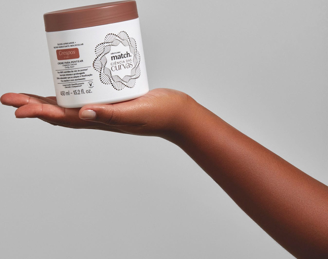 Imagem de uma mão segurando um pote branco com tampa marrom contendo creme para pentear da linha Match. Ciência das Curvas Crespos em fundo cinza.