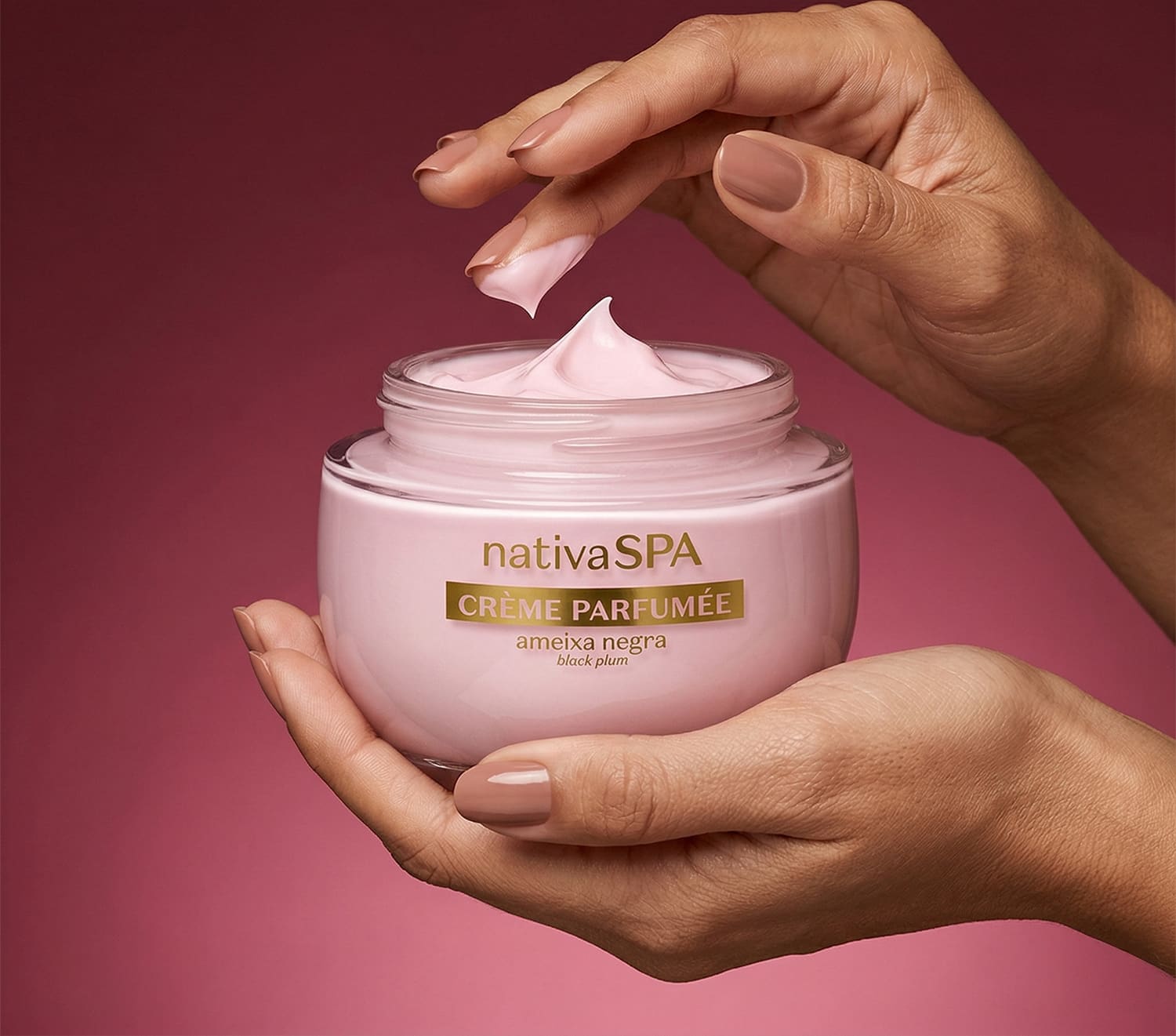 Pote aberto de creme perfumado da marca Nativa SPA Ameixa Negra repousa em mão esquerda feminina enquanto a mão direita repousa acima da embalagem aberta com um pouco de creme na ponta do dedo médio em fundo rosa escuro.