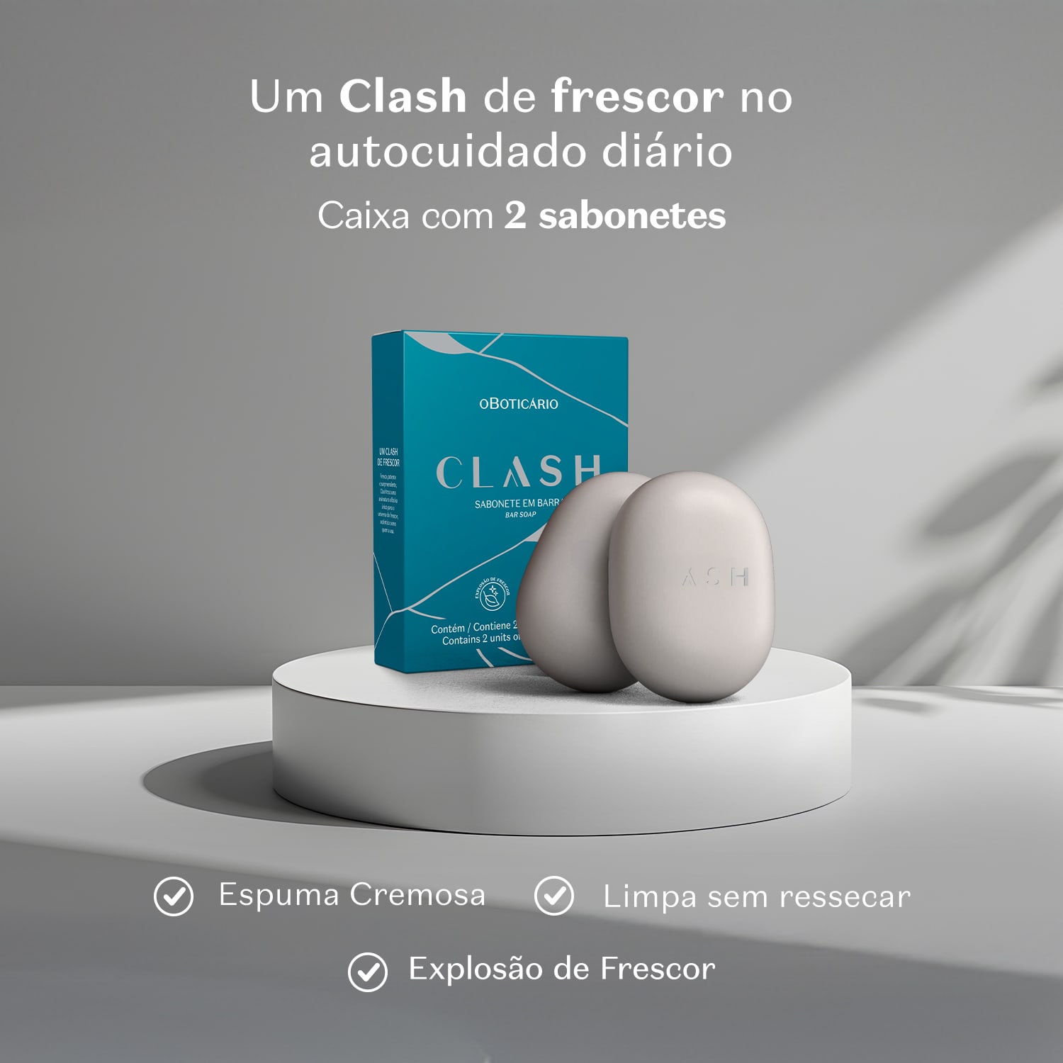 Caixa de sabonete em barra da marca Clash sobre superfície branca, com dois sabonetes brancos ao lado. Ambiente monocromático em tons de cinza. Texto informa que o produto oferece espuma cremosa, limpa sem ressecar e proporciona explosão de frescor.
