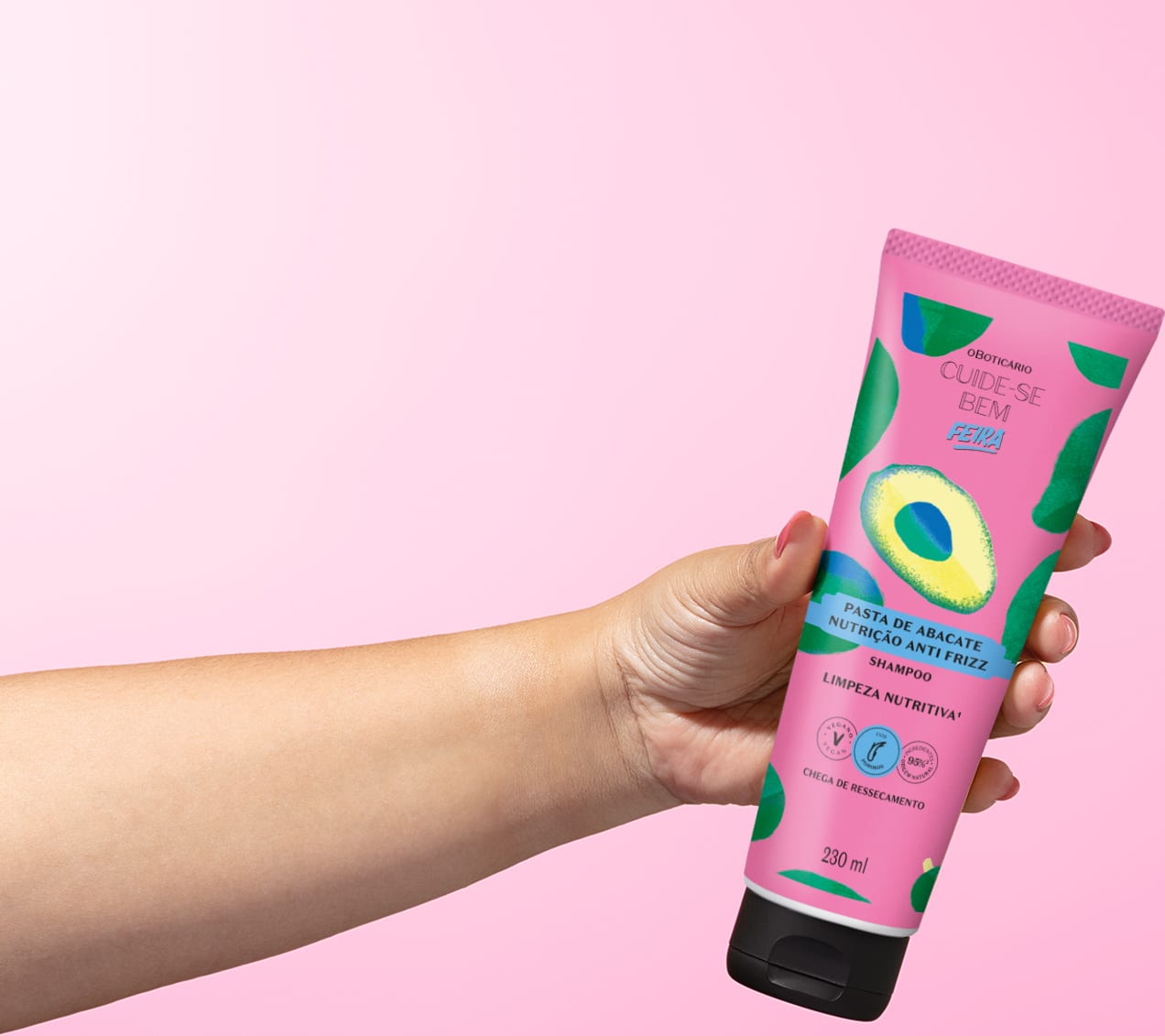 Imagem de um tubo roxo com ilustrações de abacate de shampoo da marca Cuide-se Bem Feira Pasta de Abacate, seguro por mão em fundo rosa.