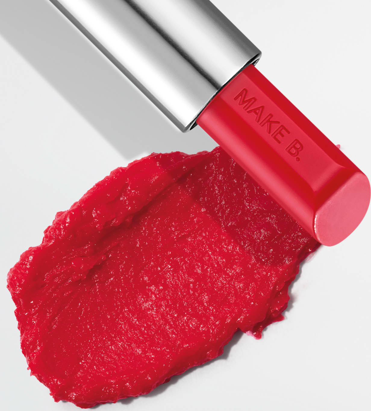 O balm labial vermelho da marca Make B. Lip Chronology Color Repair possui uma embalagem metálica prateada e produto vermelho brilhante. Há uma mancha do item sobre fundo branco, indicando a textura e a intensidade do produto.
