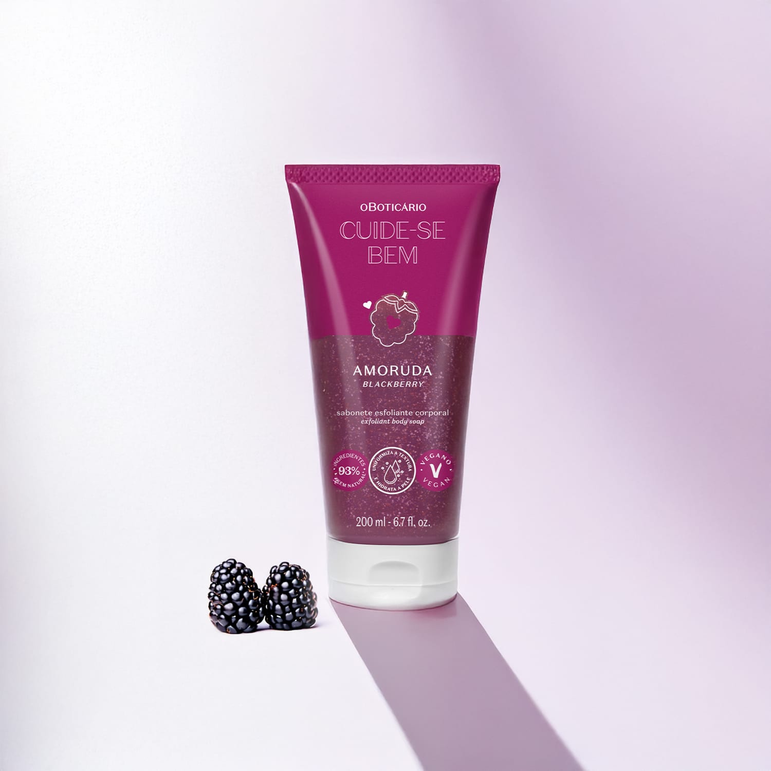 Tubo de sabonete esfoliante corporal da marca Cuide-se bem Amoruda, na cor roxa e ilustração de amora. Ao lado da embalagem há duas amoras frescas em um fundo claro com sombras.