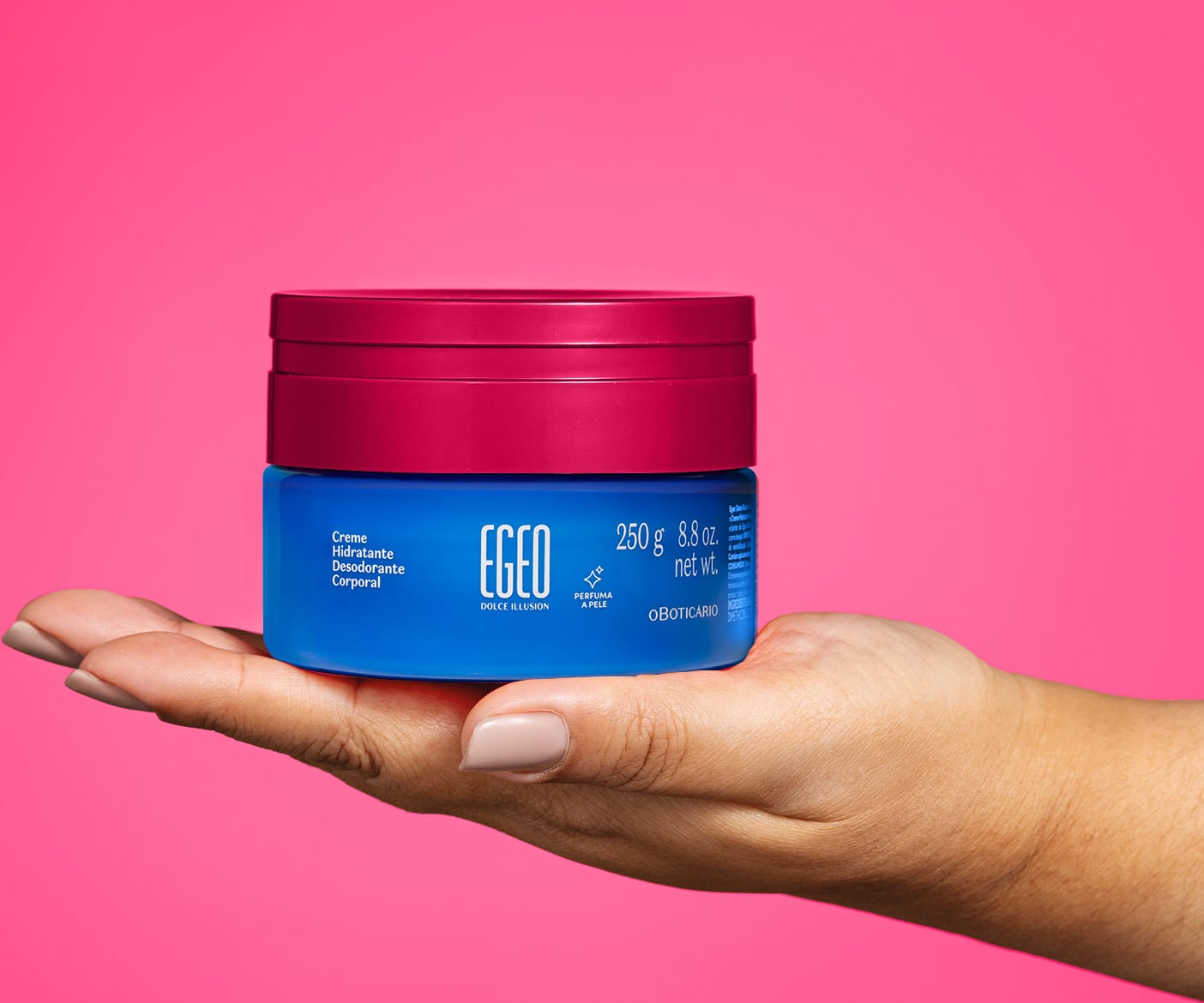 Imagem de uma mão segurando um pote azul com tampa rosa contendo 250g de creme hidratante desodorante corporal da marca Egeo Dolce Illusion. O fundo da imagem é na cor rosa liso.