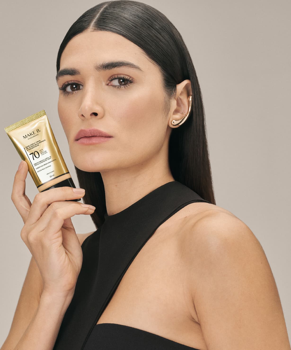 A modelo segura um tubo dourado de base líquida com protetor solar da marca Make B., em um ambiente minimalista e neutro, destacando o produto de beleza.