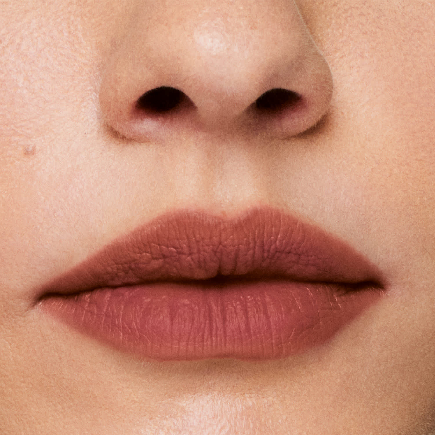 Boca feminina de mulher branca em close mostrando queixo e um pedaço do nariz, com lábios fechados com batom marrom claro sobre eles.