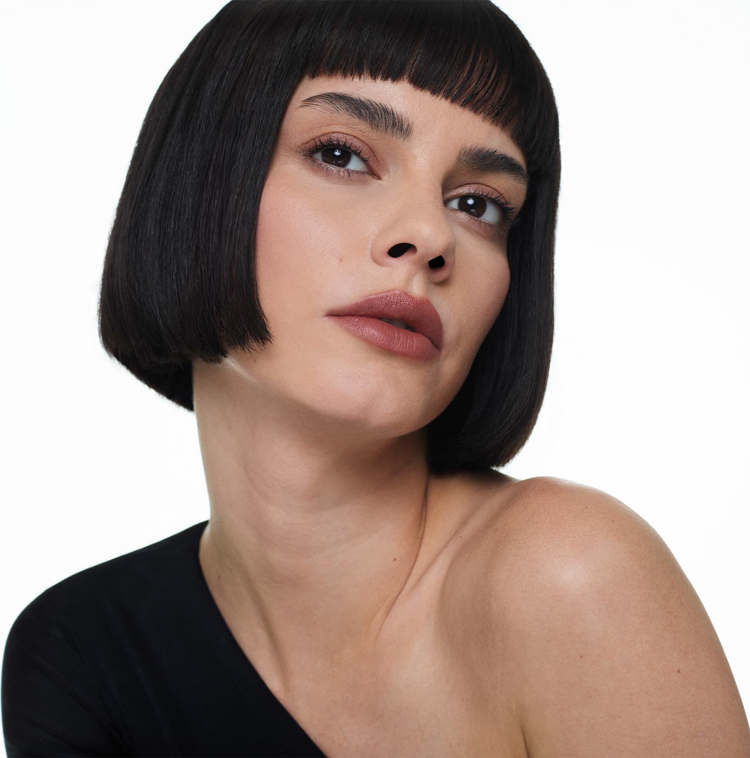Mulher branca de cabelo preto curto e franja, com expressão séria, usando roupa preta, em fundo branco om olhar sério e lábios levemente entreabertos pintados com batom marrom claro.