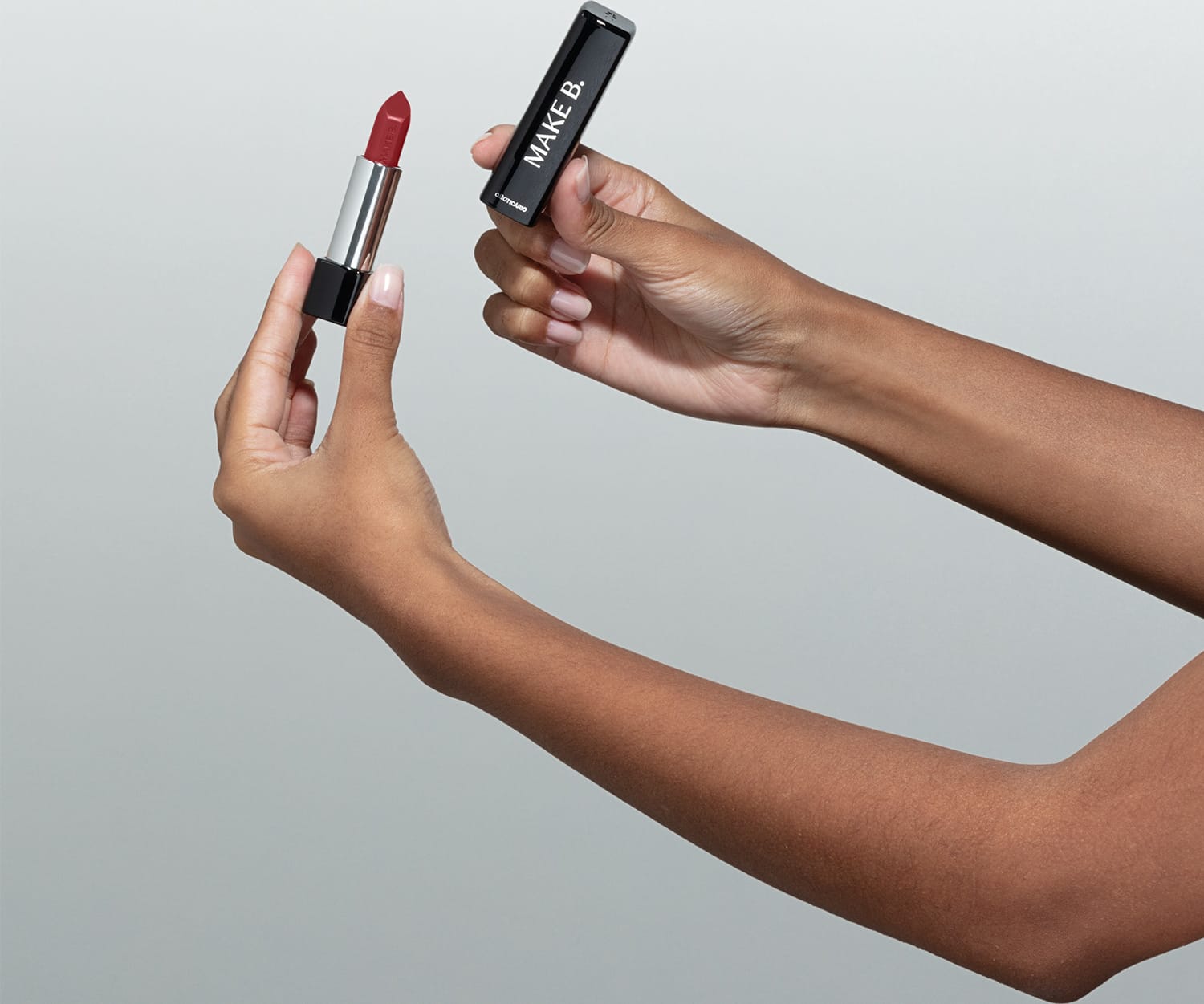 Mãos femininas segurando a tampa preta de um batom da marca Make B. na mão direita e a base do batom com a bala aparente na mão esquerda, em um ambiente minimalista e claro.