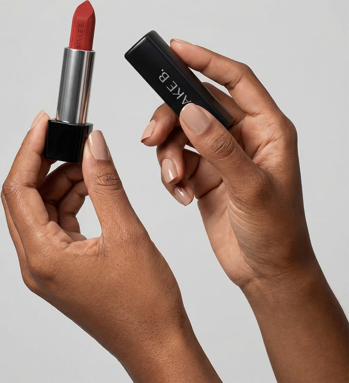 Mãos femininas segurando a tampa preta de um batom da marca Make B. na mão direita e a base do batom com a bala aparente na mão esquerda, em um ambiente minimalista e claro.