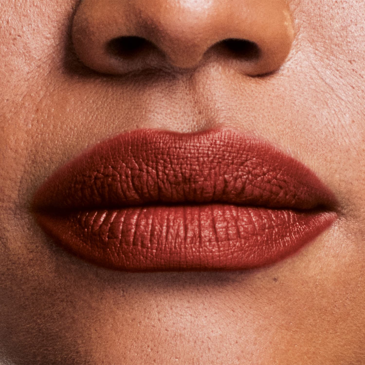 Boca feminina de mulher branca em close mostrando queixo e um pedaço do nariz, com lábios fechados com batom ocre avermelhado sobre eles.