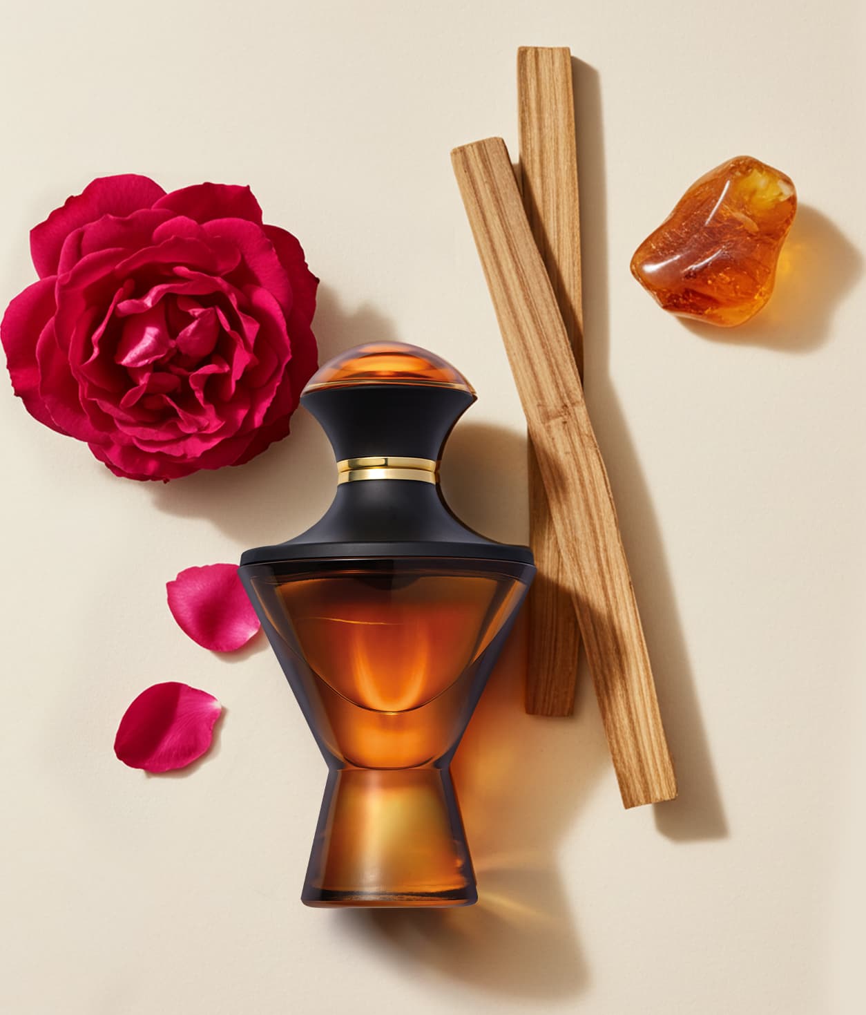 Imagem de um vidro preto e dourado de Eau de Parfum da marca Hadiya rodeado por uma rosa vermelha, cristal âmbar e varetas de madeira sobre superfície bege.