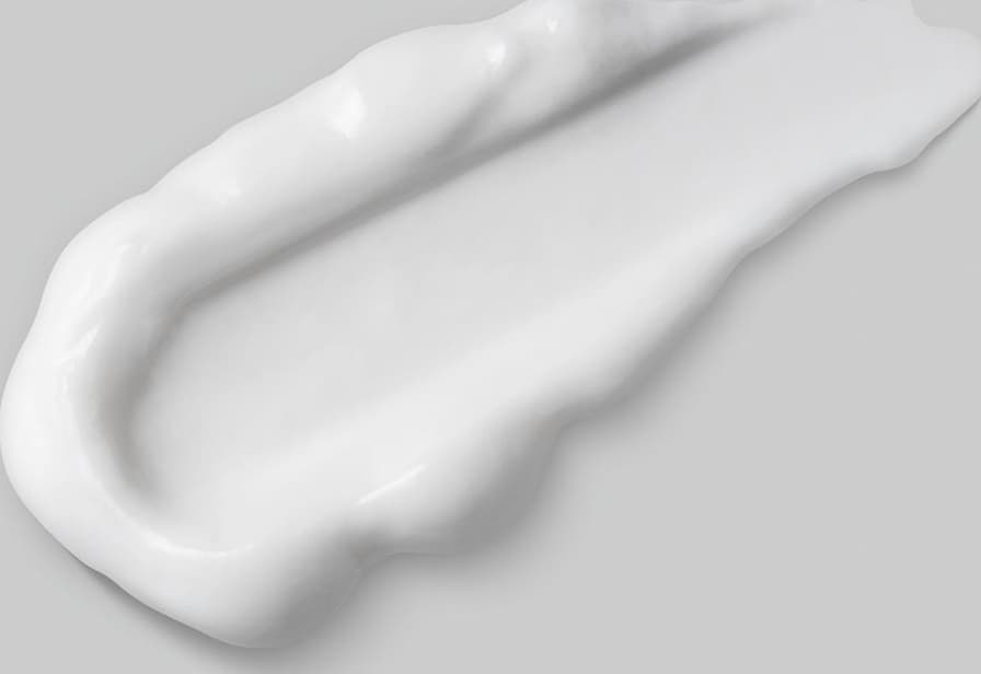 Imagem de um pouco de condicionador sobre superfície clara, lisa e uniforme, evidenciando a textura cremosa na cor branca do produto, que tem aparência suave e brilhante,