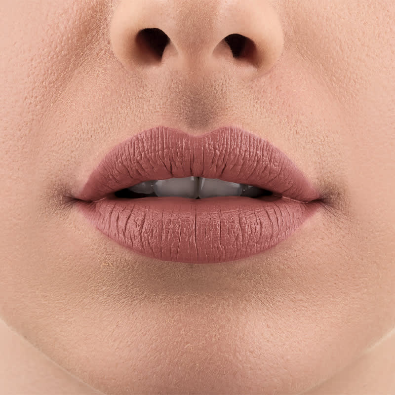 Imagem de um rosto feminino em close-up, evidenciando os lábios com o batom líquido Make B. na cor rosa. A boca está entreaberta mostrando parcialmente os dentes brancos.