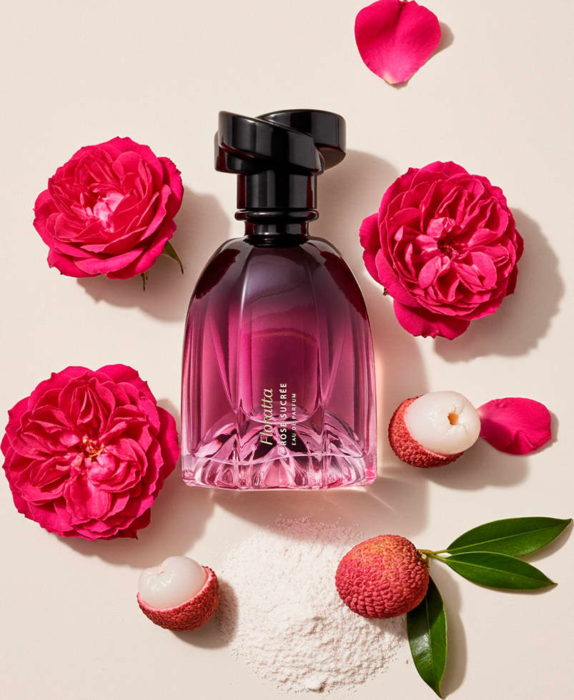 A imagem mostra um frasco de item de perfumaria Floratta Rose Sucrée em tom de roxo, rodeado por pétalas de rosas vermelhas, frutas lichia e folhas verdes sobre superfície clara.