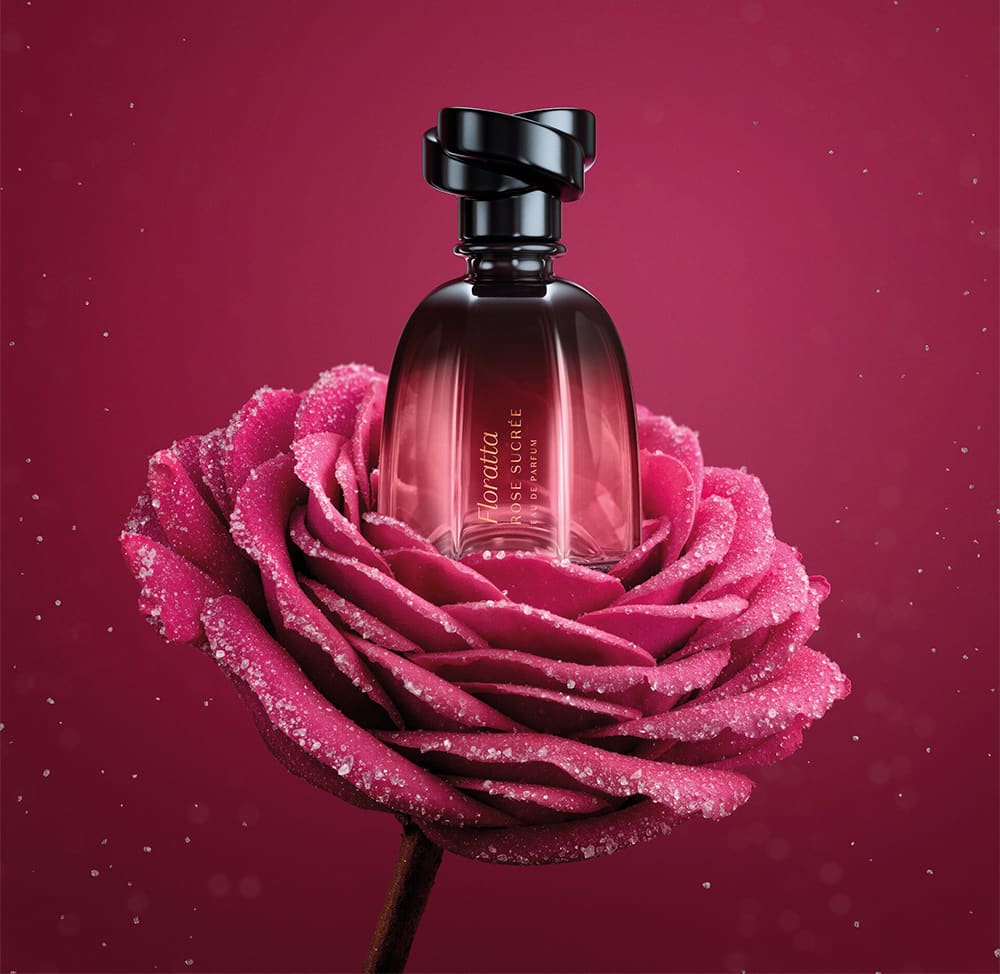 Frasco de item de perfumaria Floratta Rose Sucrée em cima de uma rosa sobre fundo vermelho-escuro. As pétalas são rosa brilhantes e possuem gotículas de orvalho.