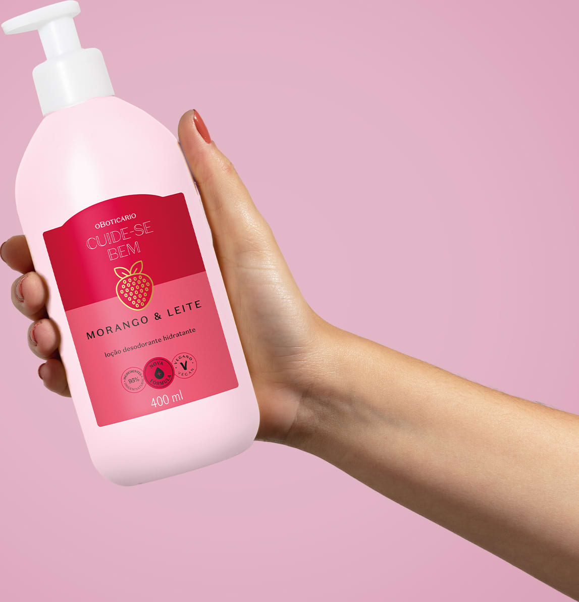Imagem de uma embalagem branca de loção hidratante corporal Cuide-se Bem Morango e Leite com rótulo vermelho e rosa e imagem de morango, segura por mão feminina contra fundo rosa.