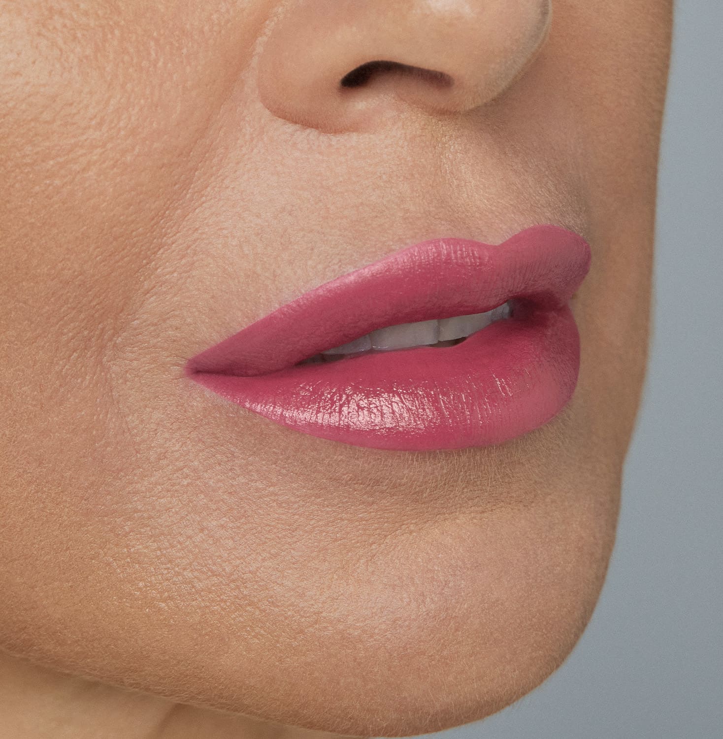 Boca feminina de mulher branca em close mostrando queixo e um pedaço do nariz, com lábios entreabertos mostrando ligeiramente os dentes brancos com batom rosado brilhante.