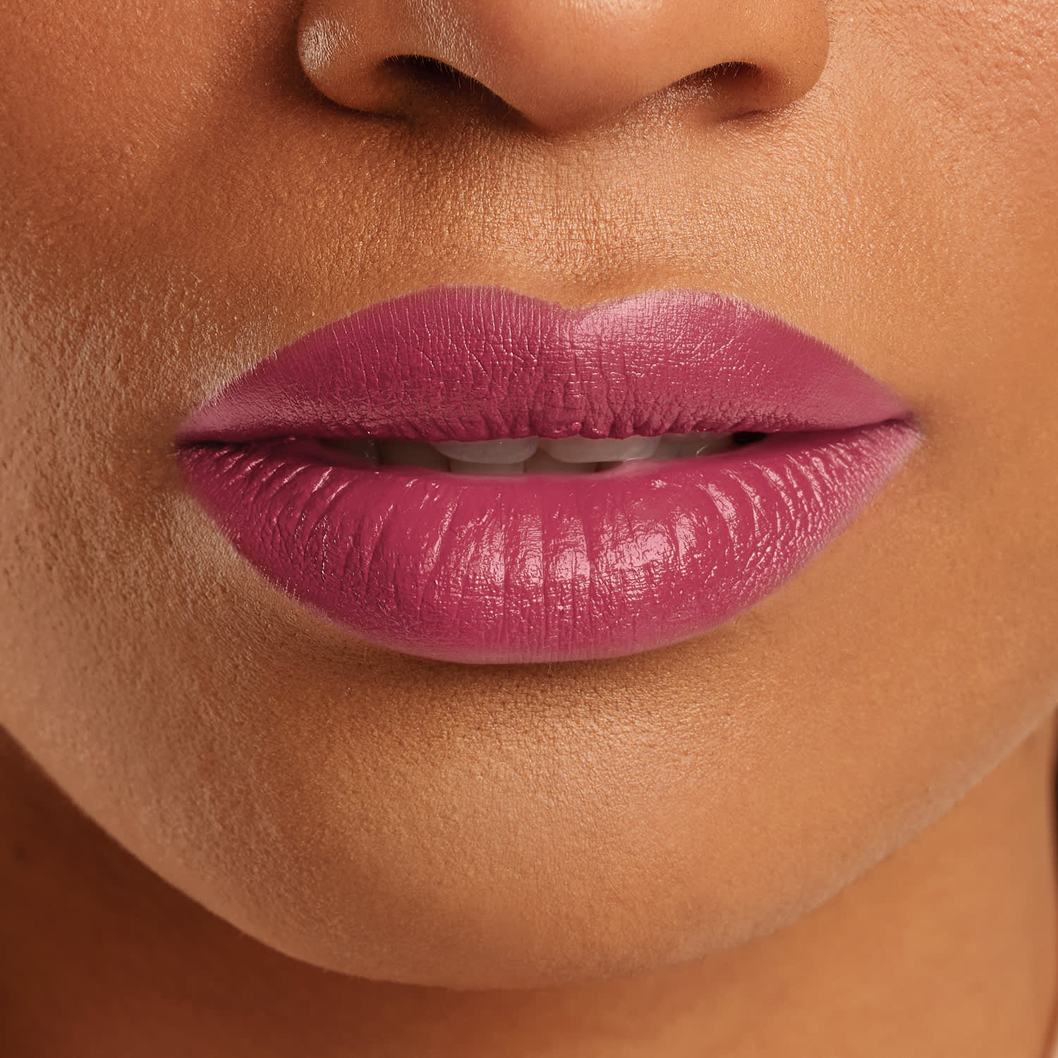 Boca feminina de mulher parda em close mostrando queixo e um pedaço do nariz, com lábios entreabertos mostrando ligeiramente os dentes brancos com batom rosado brilhante.