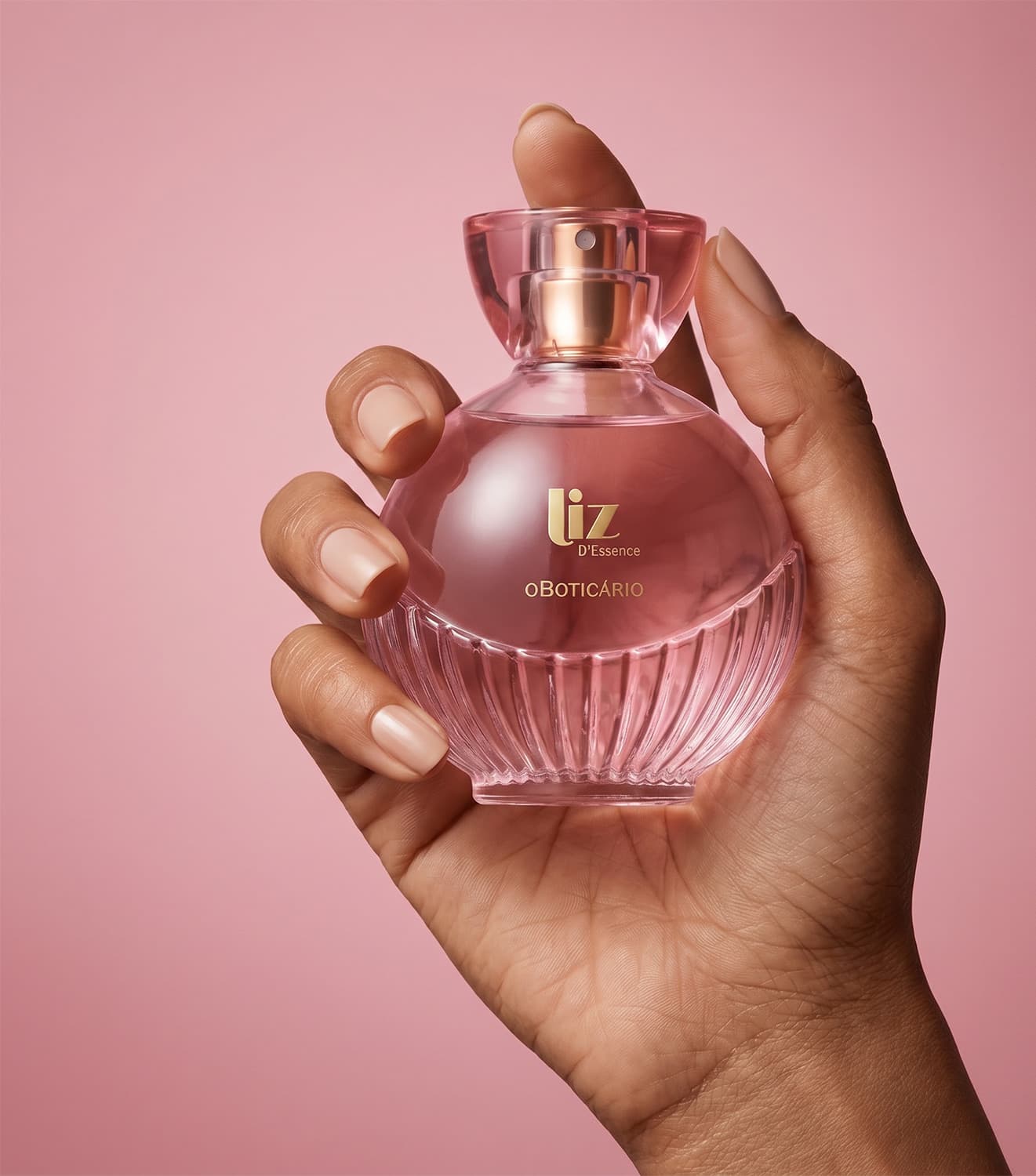 Imagem de um frasco de perfume rosa da marca Liz d'Essence, em uma mão feminina, em um fundo rosa sólido, sugerindo um ambiente de beleza e cosmética.