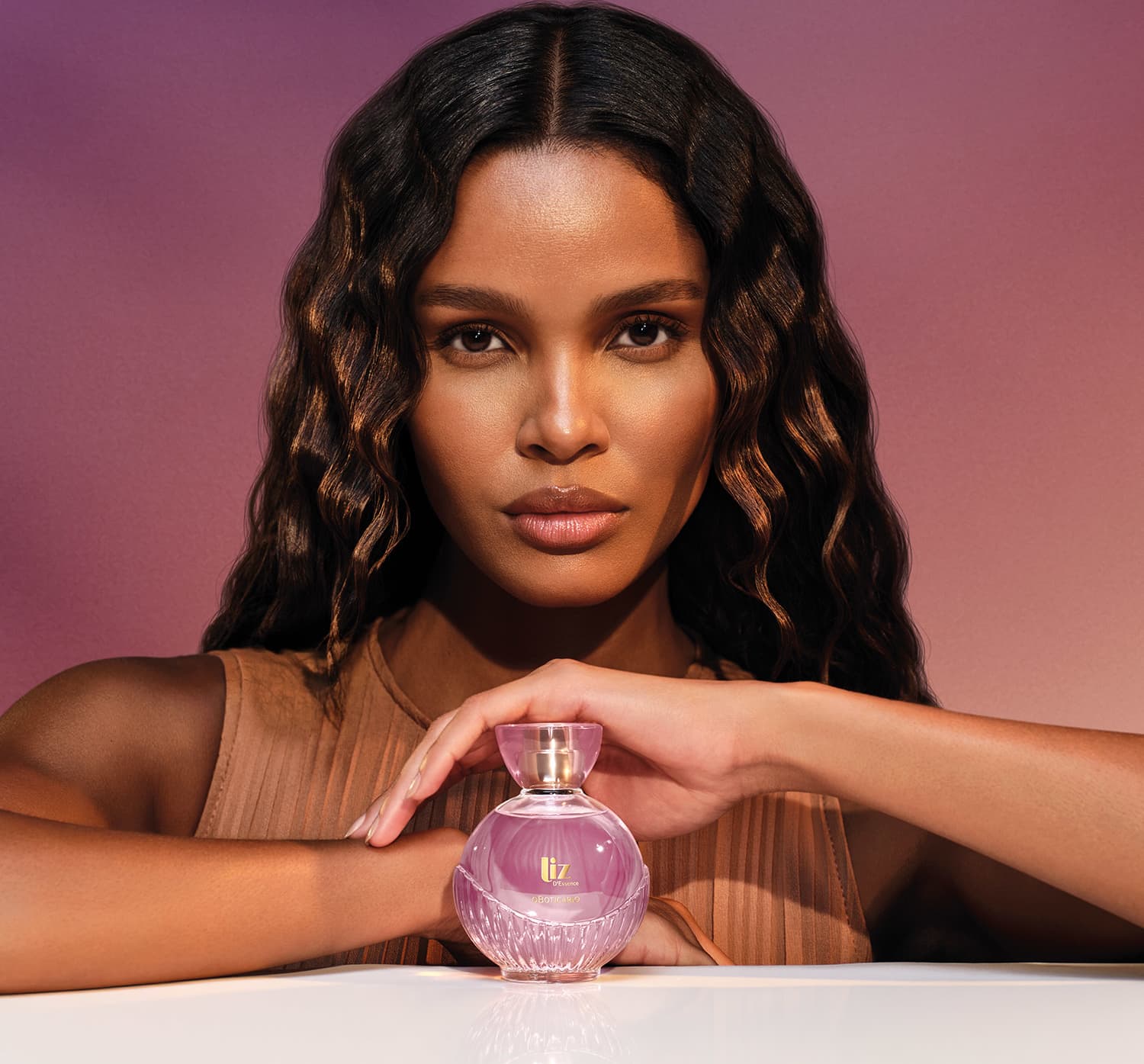 Modelo negra com cabelos longos e ondulados e expressão séria vista dos ombros para cima. A frente dela, um frasco de desodorante colônia da marca Liz D'Essence, sobre o qual ela repousa sua mão esquerda.
