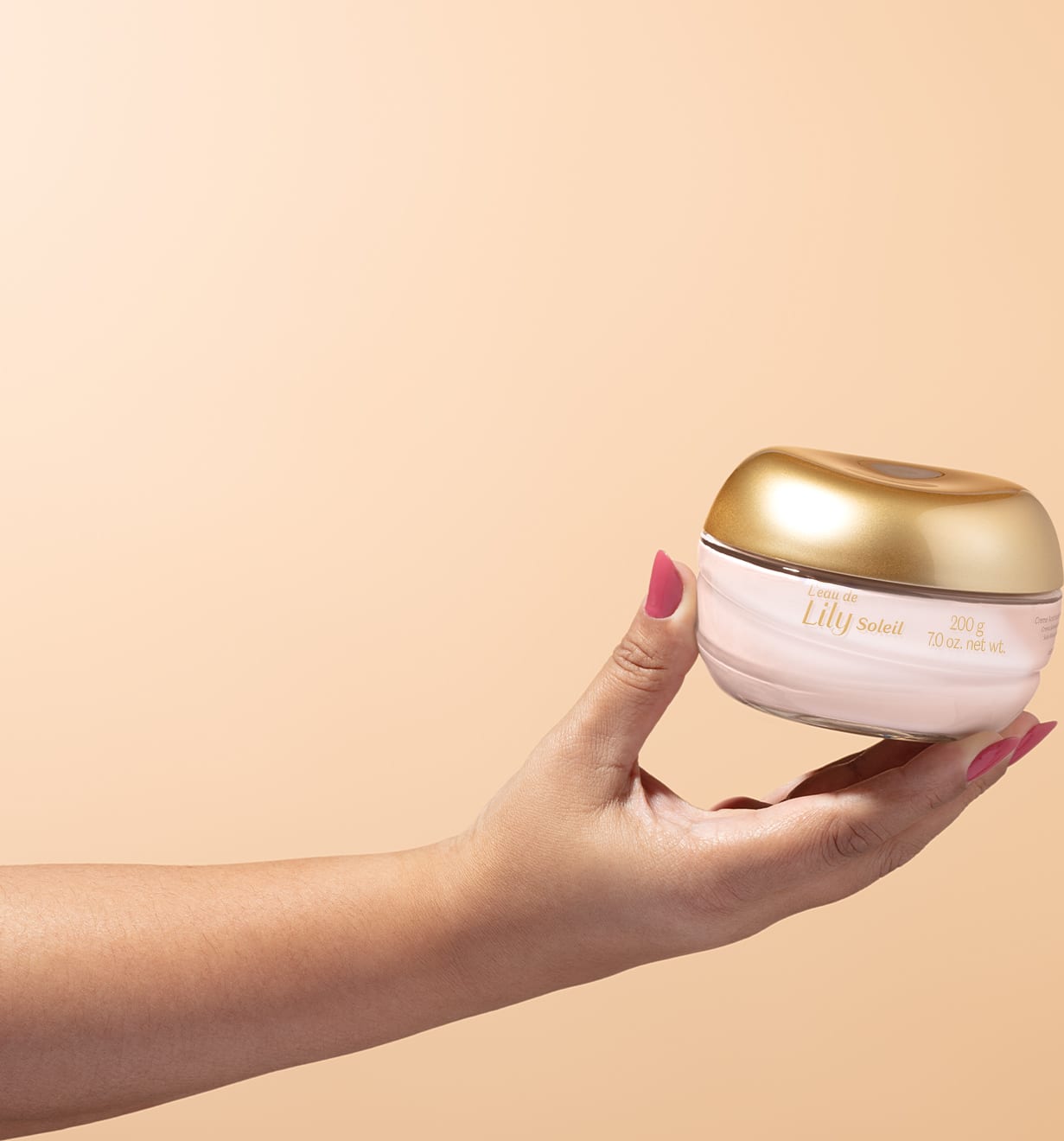 Imagem de uma mão feminina segurando um pote de vidro transparente com conteúdo branco e tampa dourada contendo creme acetinado corporal da marca L'eau de Lily Soleil em fundo bege vibrante.