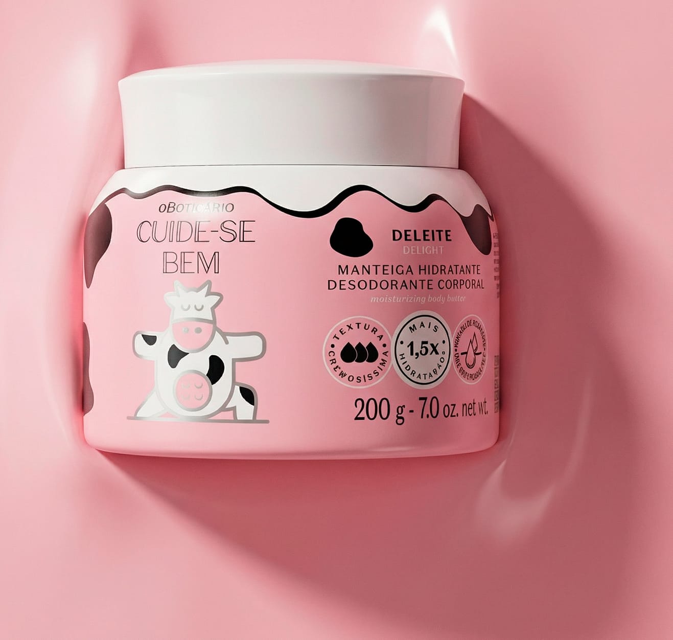 A imagem mostra o pote branco de manteiga corporal Cuide-se Bem Deleite deitado em uma textura cremosa na cor rosa. A embalagem é branca com rótulo em rosa e estampa de vaca. Ele tem tampa branca e contém 200g de produto.