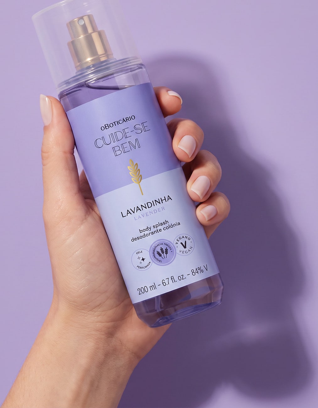 Frasco de item de perfumaria da marca Cuide-se bem, na cor lilás, sendo segurado por mão feminina sobre fundo liso e monocromático na mesma tonalidade.