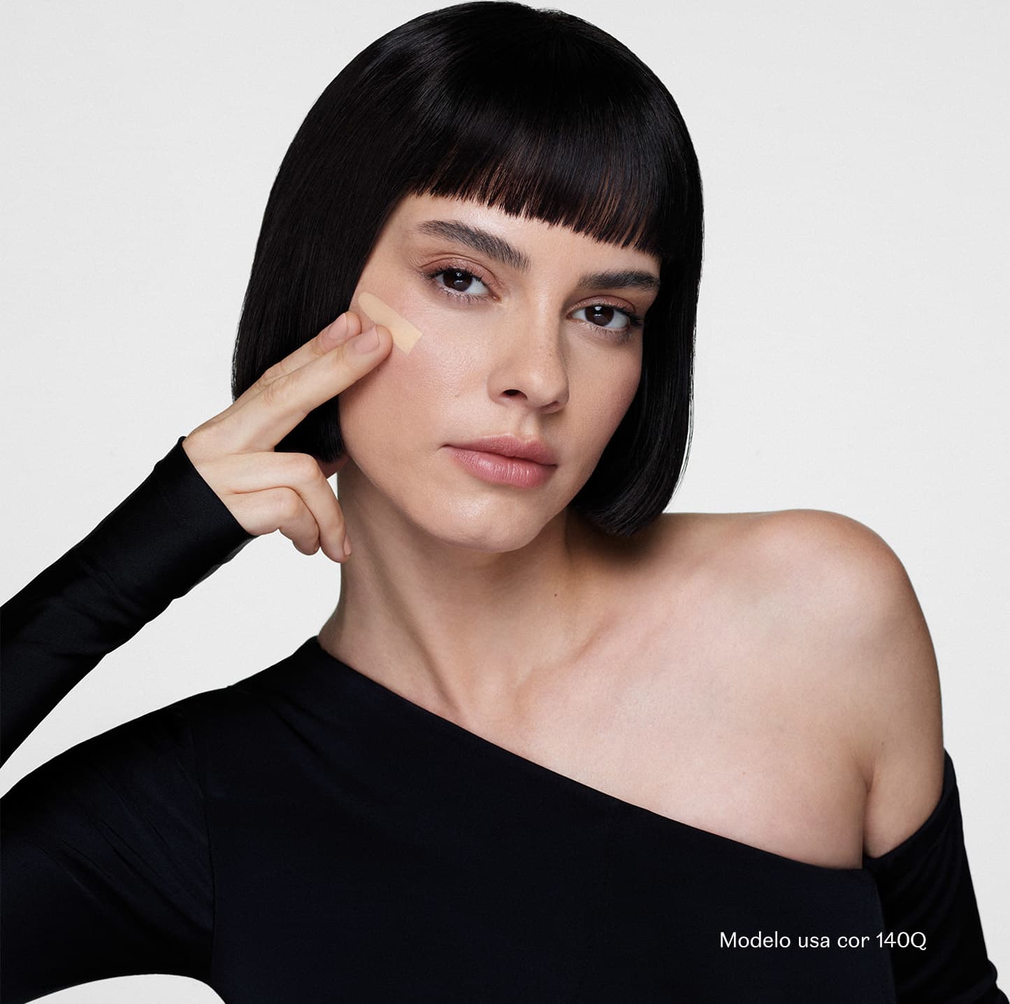 Mulher branca de cabelo preto curto e franja, com expressão séria, usando roupa preta, em fundo branco. Com o indicador e o dedo médio da mão direita, ela aplica um pouco de base clara sobre a maçã do rosto.