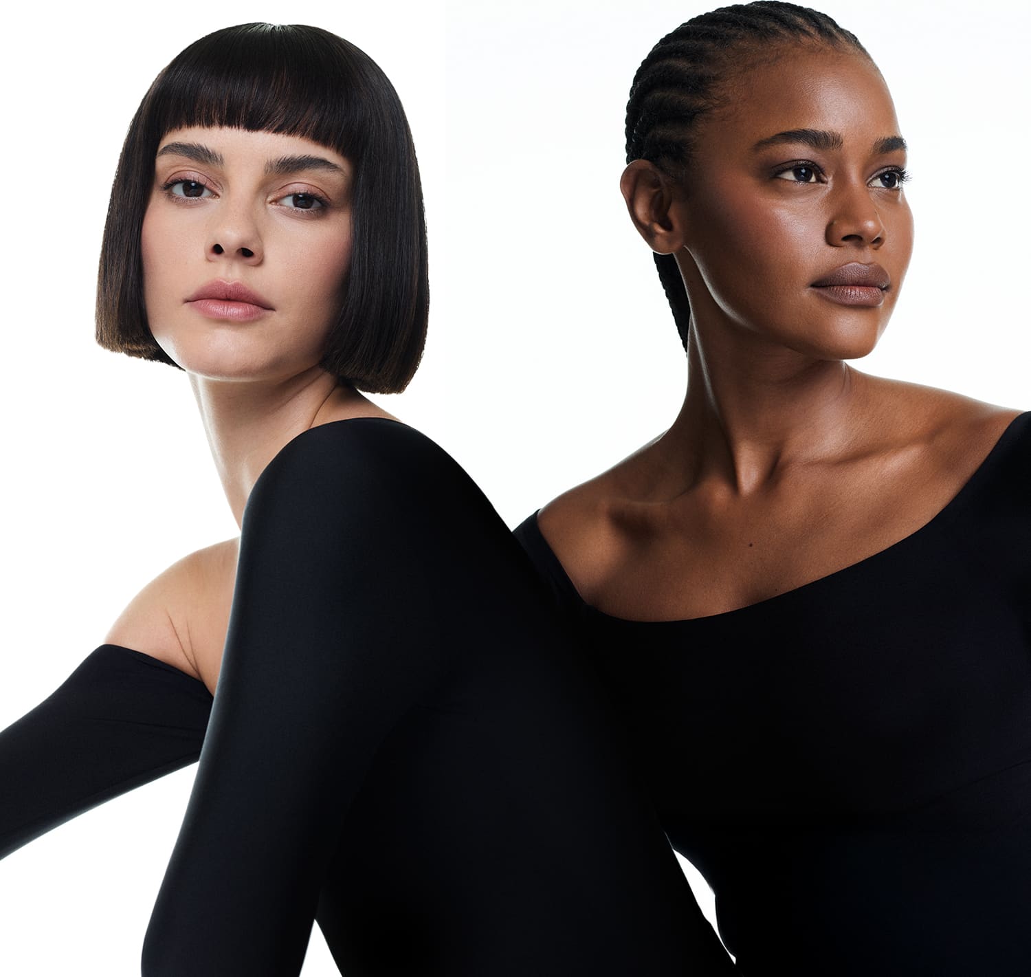 Duas modelos vestindo roupas pretas, sendo uma mulher negra com cabelo trançado preso e outra branca com cabelo curto preto e franja em pose empoderada em fundo branco.