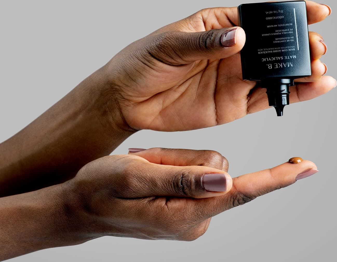 Mãos femininas negras posicionadas uma em cima da outra, sendo que a de cima segura um frasco de base Make B. aberto e na mão de baixo, vemos uma gota de base na ponta do indicador da modelo em fundo cinza claro.