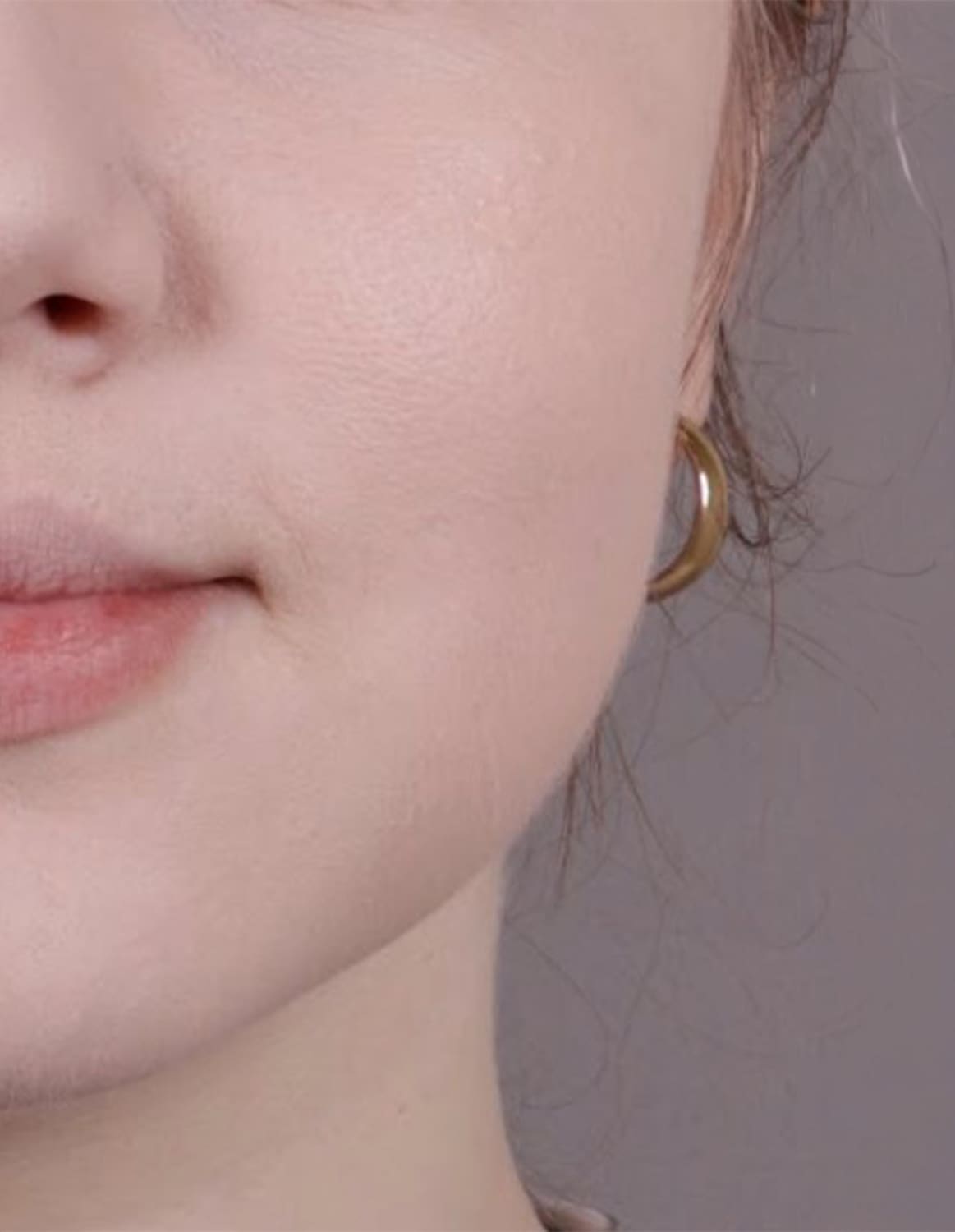 Imagem de close-up do quarto inferior esquerdo de um rosto feminino branco com mecha de cabelo loiro e curto aparente com brinco de argola pequeno dourado, em um fundo cinza claro.