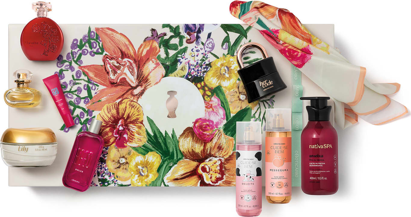 A imagem mostra uma superfície branca com arranjo de itens de perfumaria e cuidados pessoais, incluindo frascos, caixas com flores e folhagens estampadas e embalagens variadas dos produtos o Boticário.