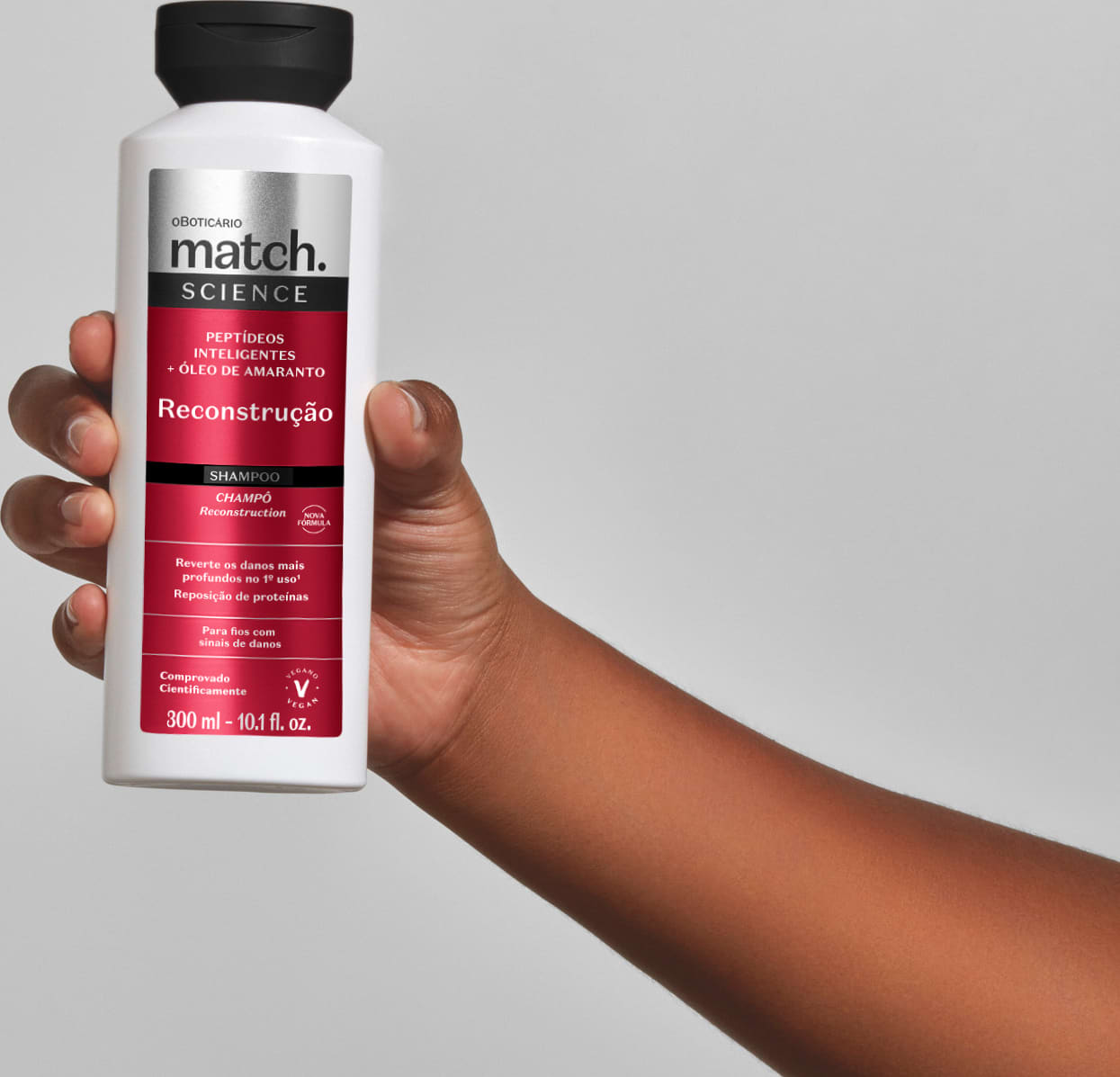 Frasco branco com rótulo vermelho da linha Match Science contendo shampoo para reconstrução capilar, seguro por uma mão feminina em fundo cinza claro.