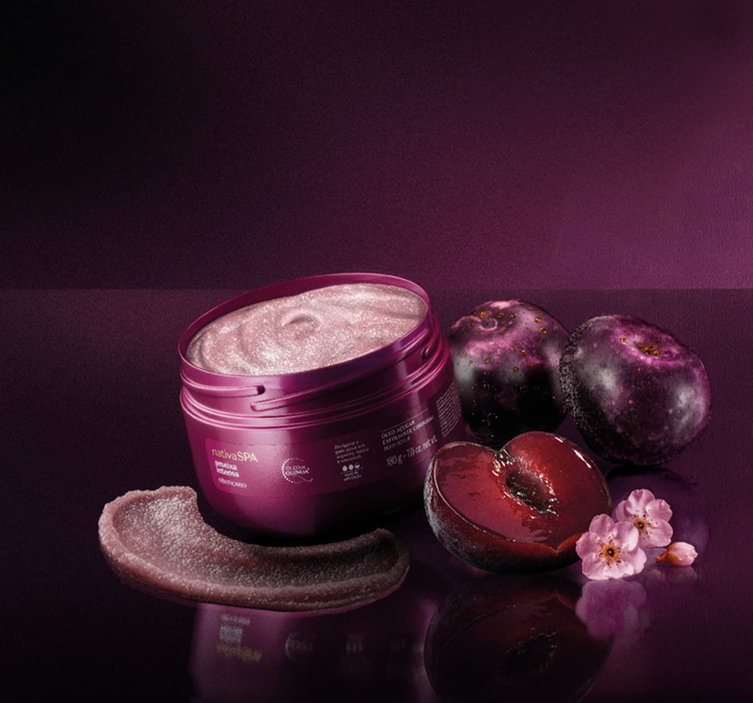Pote de creme cosmético rosa em superfície escura, com flores e frutos roxos ao redor, em ambiente de penumbra com tons de roxo e preto.