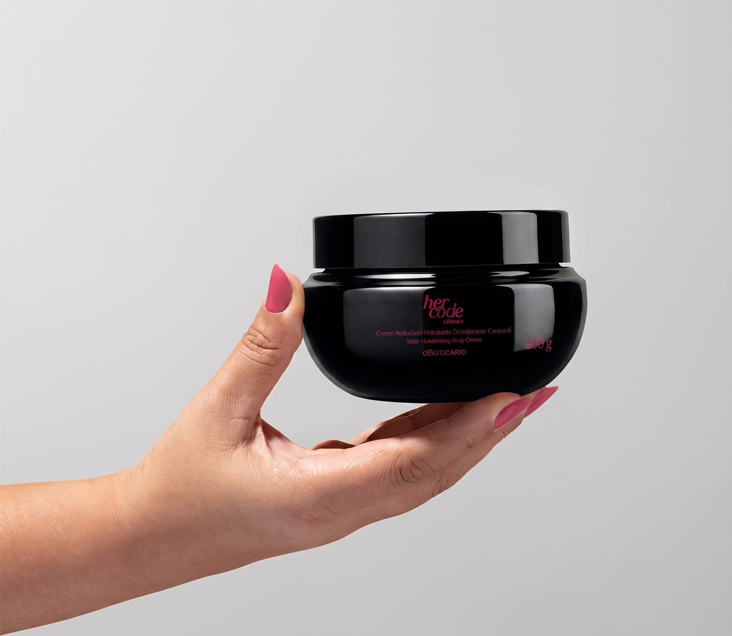 Recipiente preto de item de corpo e banho da marca Her Code, seguro por mão feminina com unhas pintadas de rosa, sobre fundo branco liso.