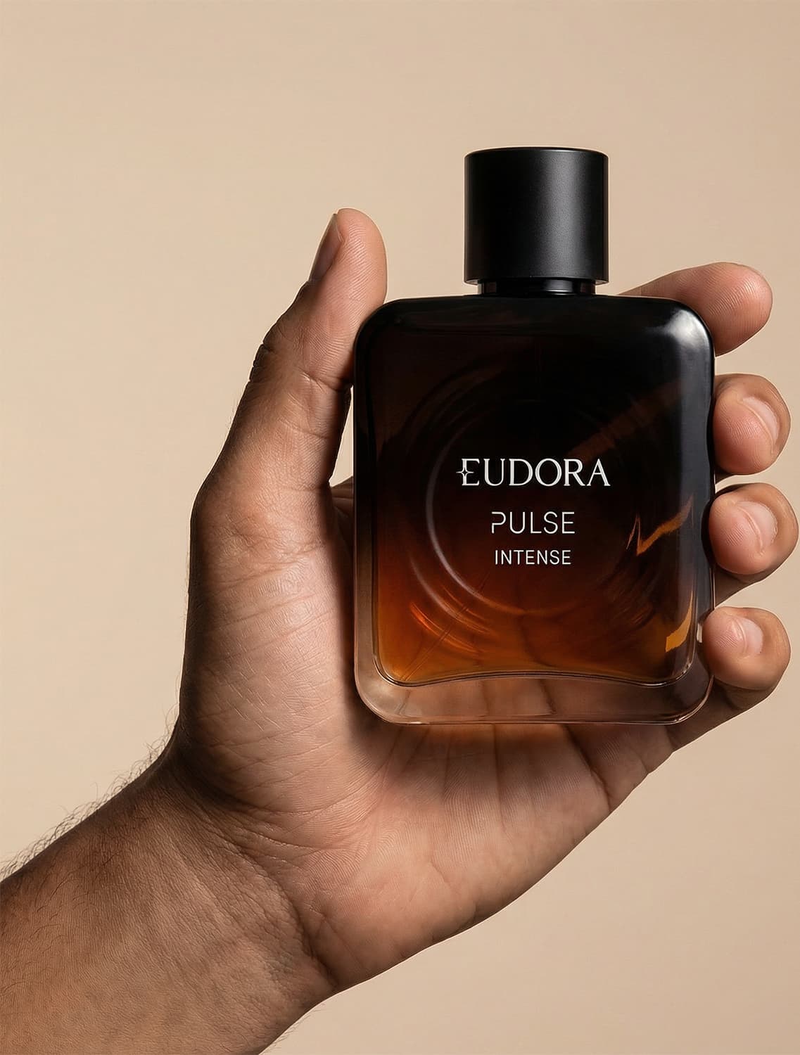 Imagem de um frasco do desodorante colônia da marca Eudora Pulse Intense, em um fundo bege claro. O produto é segurado por uma mão masculina.