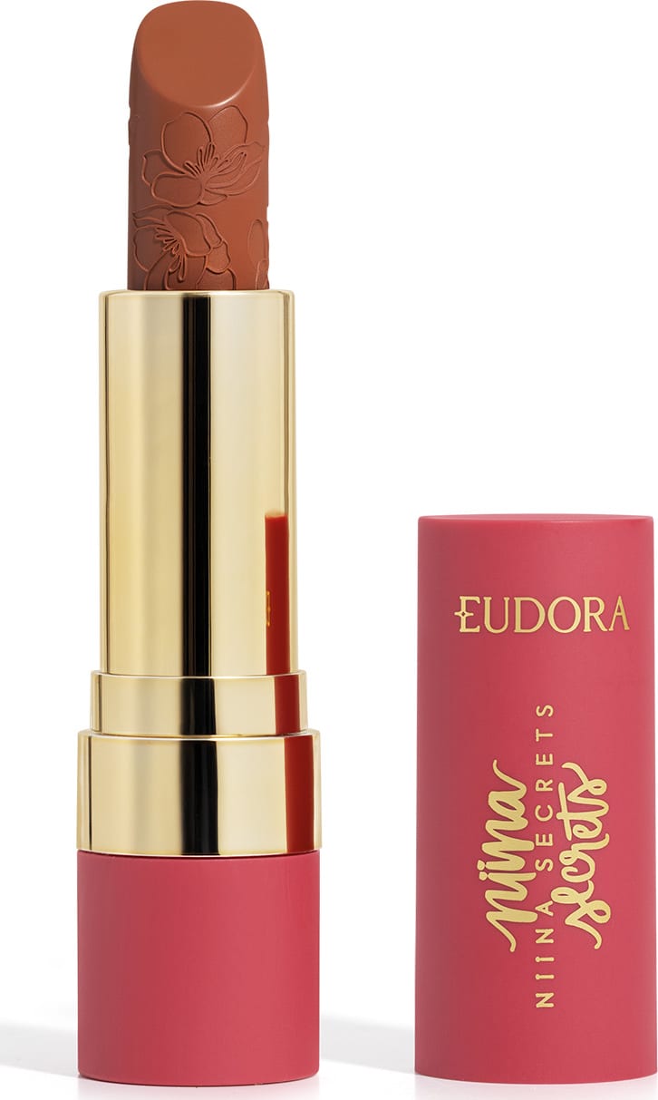 Embalagem de um batom aberto, com corpo dourado e tampa e base rosa, escrito "Eudora Niina Secrets". A cor do batom é castanho e tem um relevo exclusivo em padrão floral na bala. Produto de beleza com design elegante e sofisticado.
