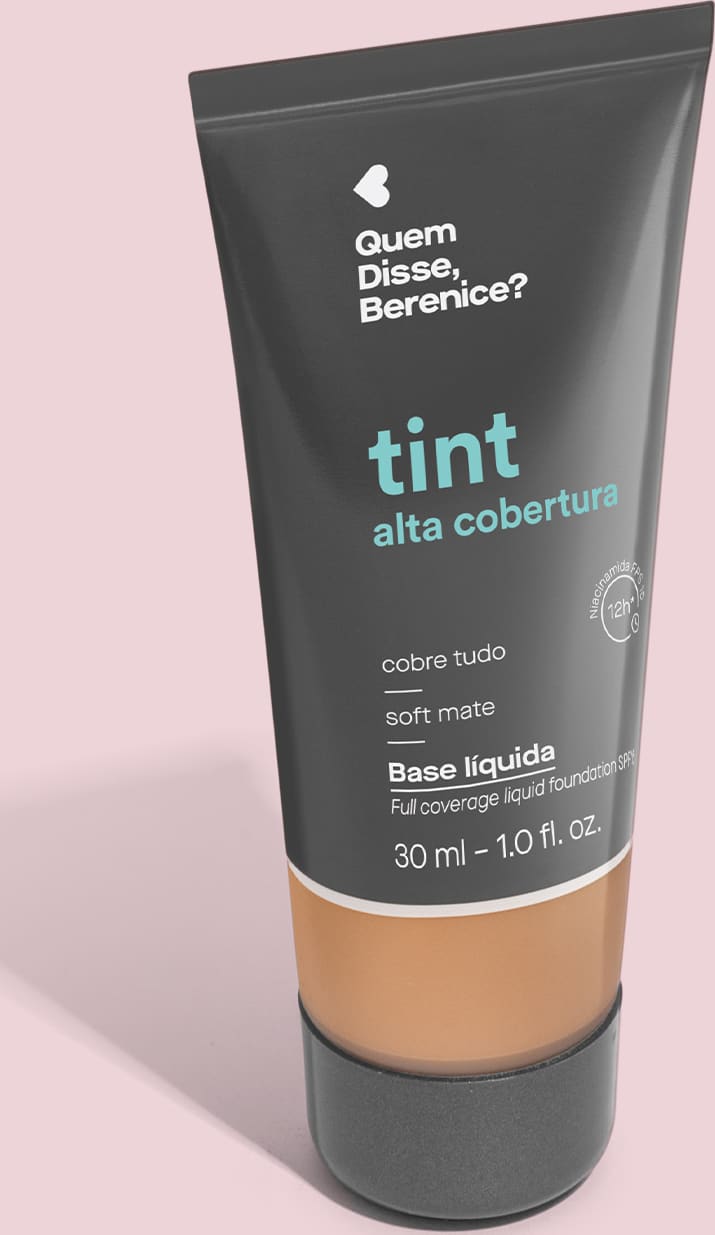 A imagem mostra a Base Tint Alta Cobertura da Quem Disse, Berenice? Embalagem cinza escura com parte inferior transparente para mostrar a cor do produto sobre fundo rosa.