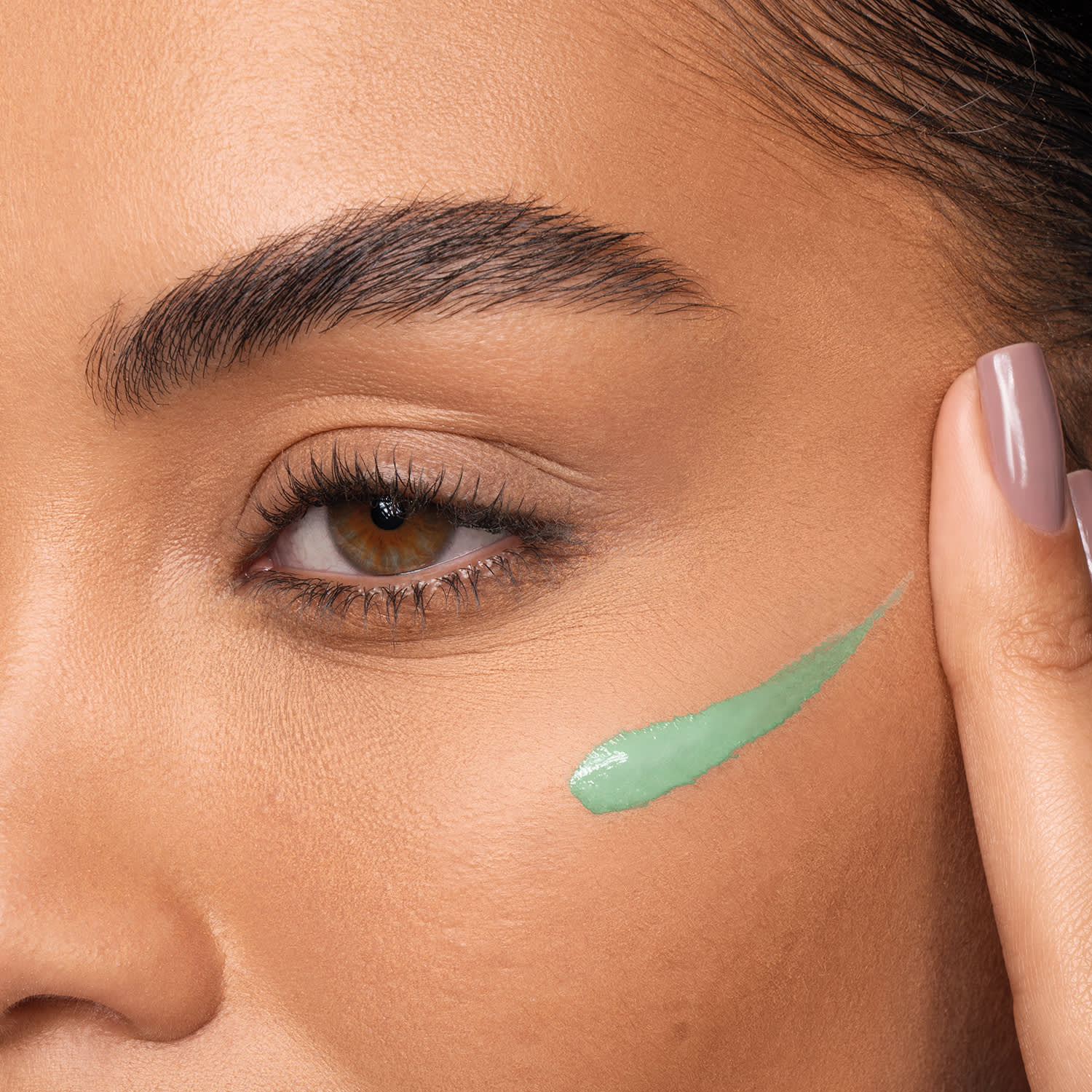 A imagem mostra uma mulher jovem aplicando uma amostra do corretivo na cor verde.