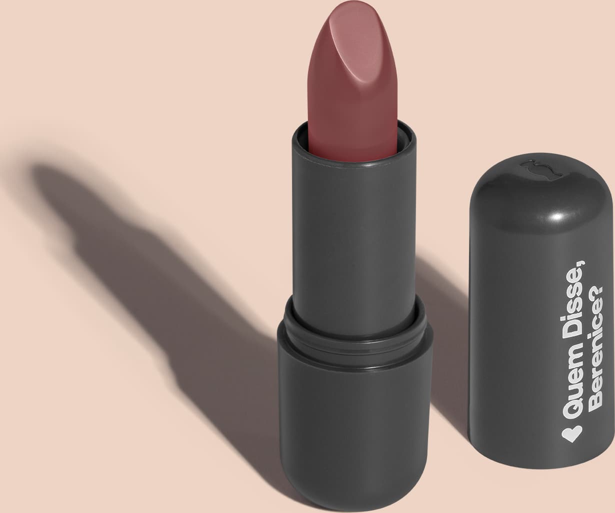 Batom de cor marrom com fundo rosado em embalagem preta com logotipo da marca Quem Disse, Berenice? em destaque. O batom está aberto sobre superfície rosa com iluminação que cria uma longa sombra projetada atrás da embalagem.