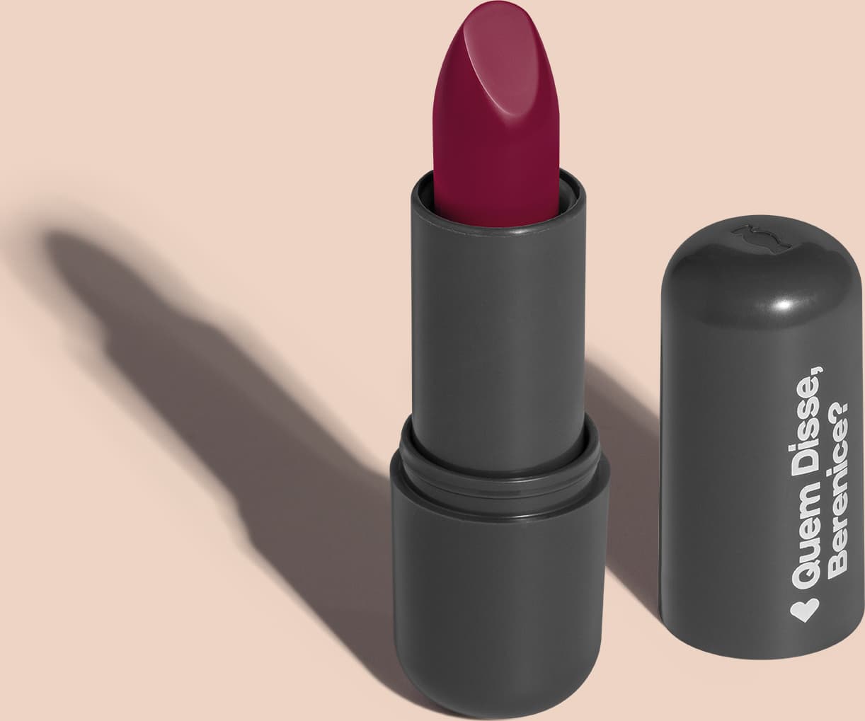Batom de cor pink em embalagem preta com logotipo da marca Quem Disse, Berenice? em destaque. O batom está aberto sobre superfície rosa com iluminação que cria uma longa sombra projetada atrás da embalagem.