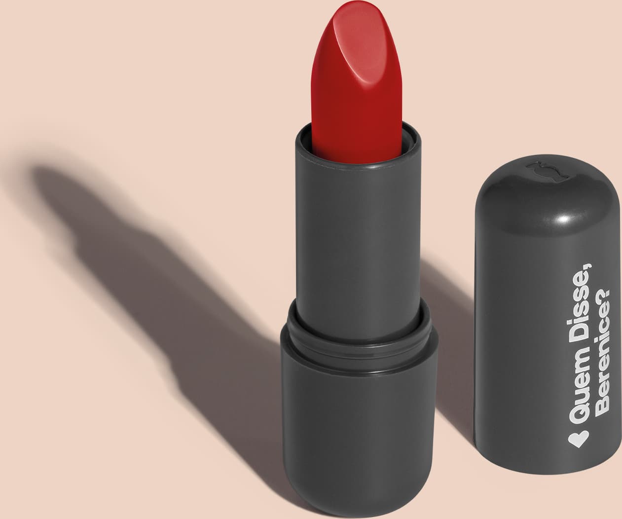 Batom de cor vermelho vibrante em embalagem preta com logotipo da marca Quem Disse, Berenice? em destaque. O batom está aberto sobre superfície rosa com iluminação que cria uma longa sombra projetada atrás da embalagem.