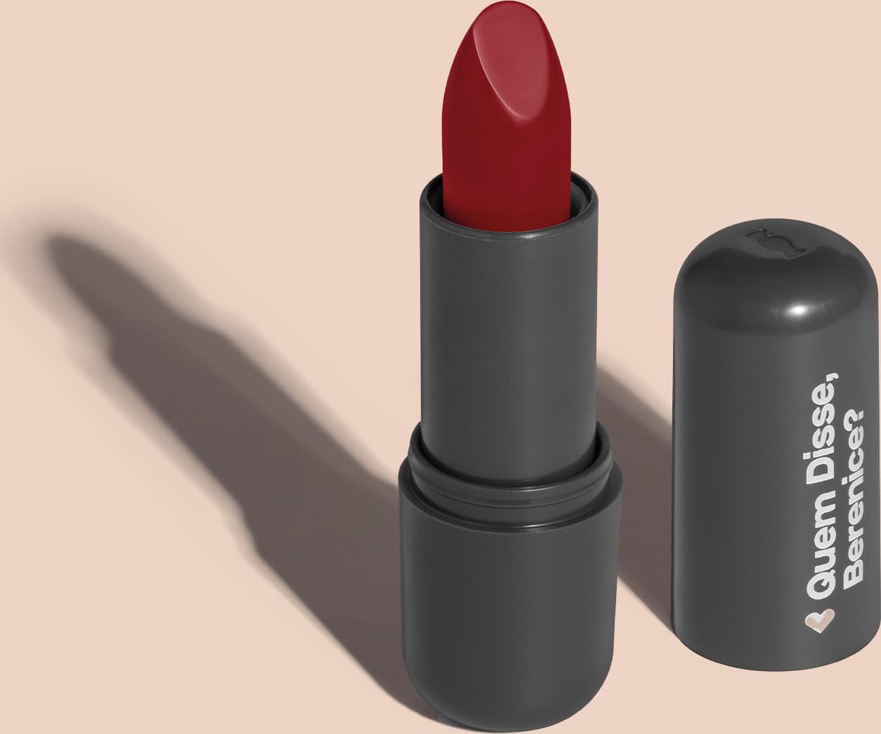 Batom de cor vermelho fechado em embalagem preta com logotipo da marca Quem Disse, Berenice? em destaque. O batom está aberto sobre superfície rosa com iluminação que cria uma longa sombra projetada atrás da embalagem.