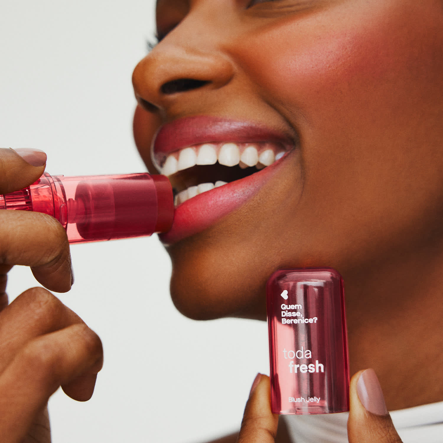 Uma mulher sorrindo passa blush jelly na cor pink da marca Toda Fresh Quem Disse, Berenice? no rosto com uma mão e a outra segura a tampa cor rosa do produto, em um fundo cinza claro.