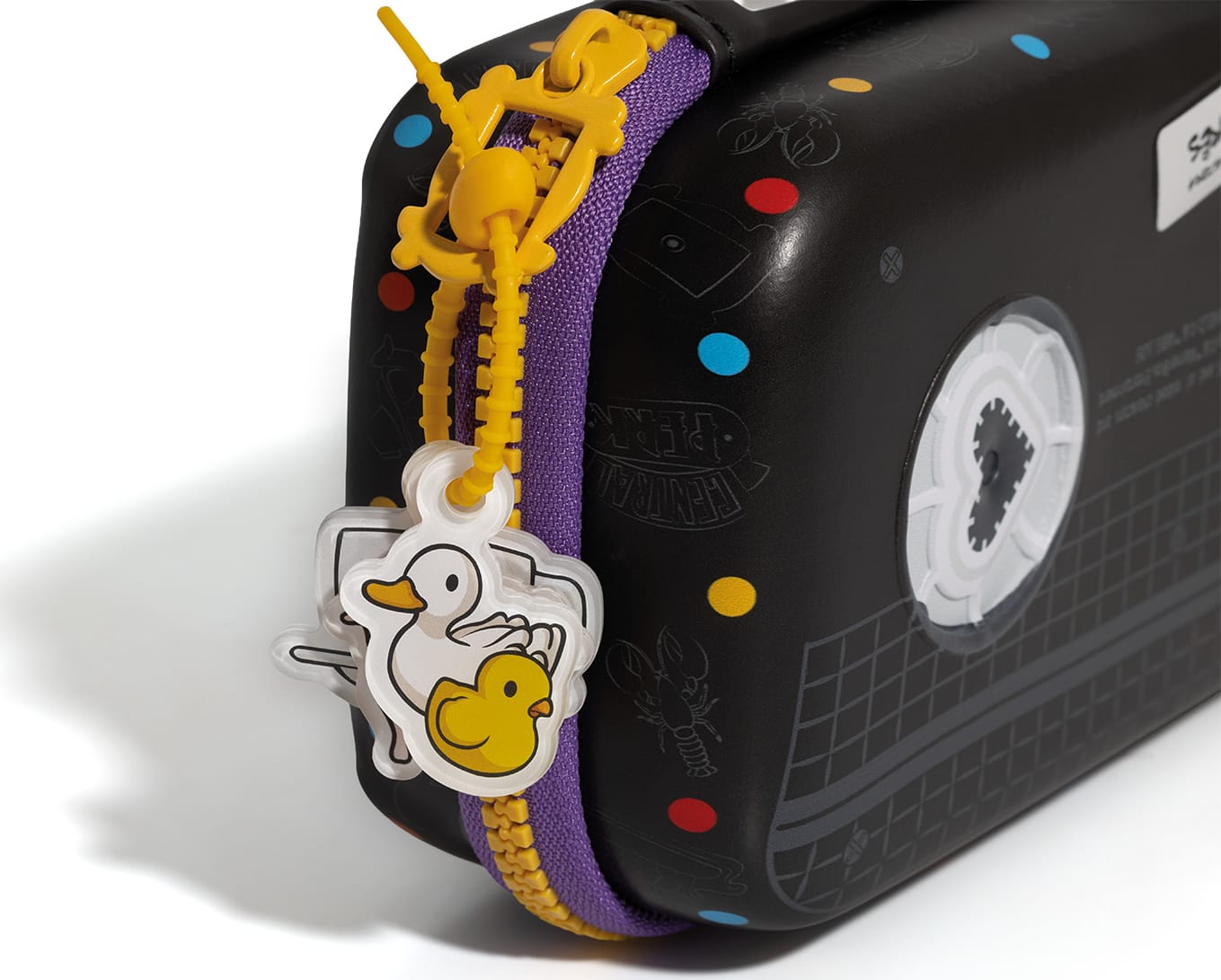 Bolsa preta com estampa de bolinhas coloridas e zíper amarelo. Pendurado no zíper há um pingente em formato de pato amarelo. 