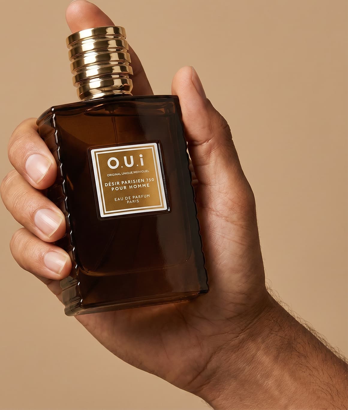 Recipiente de item de perfumaria da marca O.U.i em mãos masculinas sobre fundo bege, com detalhes dourados na embalagem.